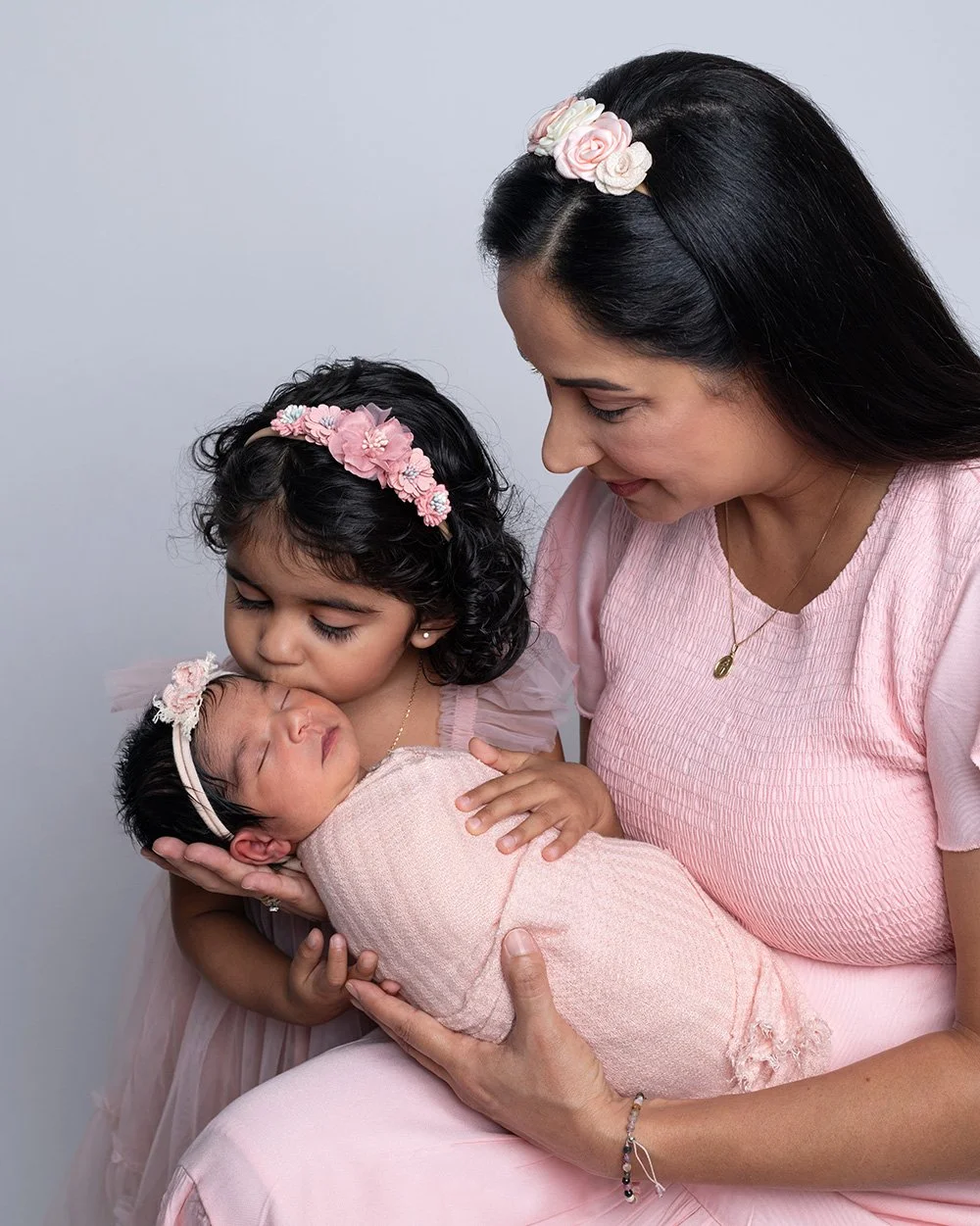 A woman and two young girls dressed in pink are holding a newborn baby wrapped in a pink blanket. The women and girls wear pink floral headbands. The woman gazes at the baby, and one girl kisses the baby's forehead.