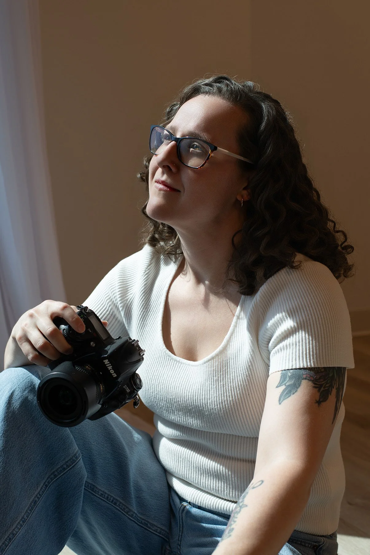 A woman with curly hair and glasses holding a Nikon camera, sitting in a relaxed pose indoors, wearing a white ribbed shirt and blue jeans, with a tattoo on her arm.