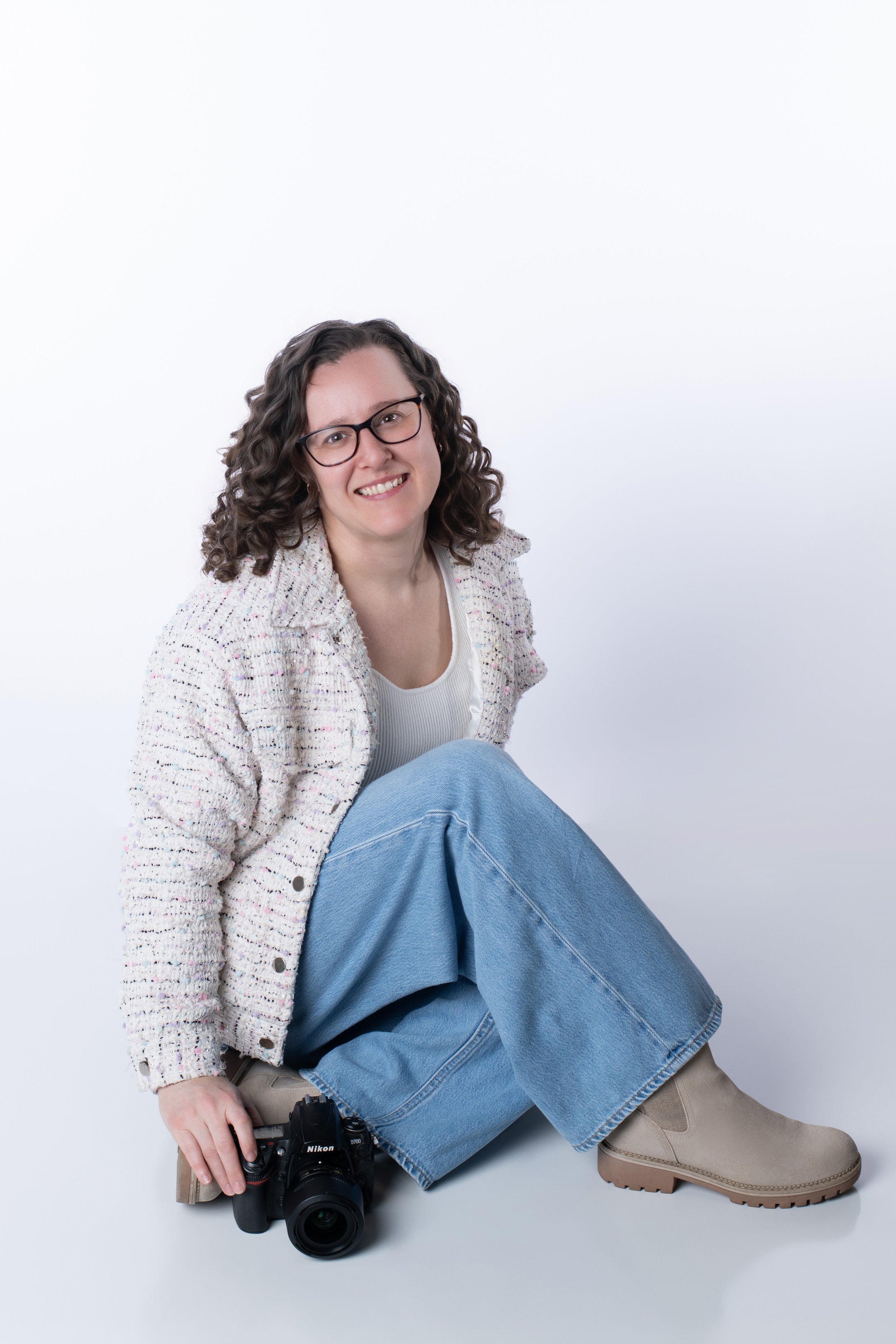 A smiling woman with curly hair, glasses, wearing a light-colored sweater, sitting on the floor with one leg bent, holding a Nikon camera, against a plain white background.