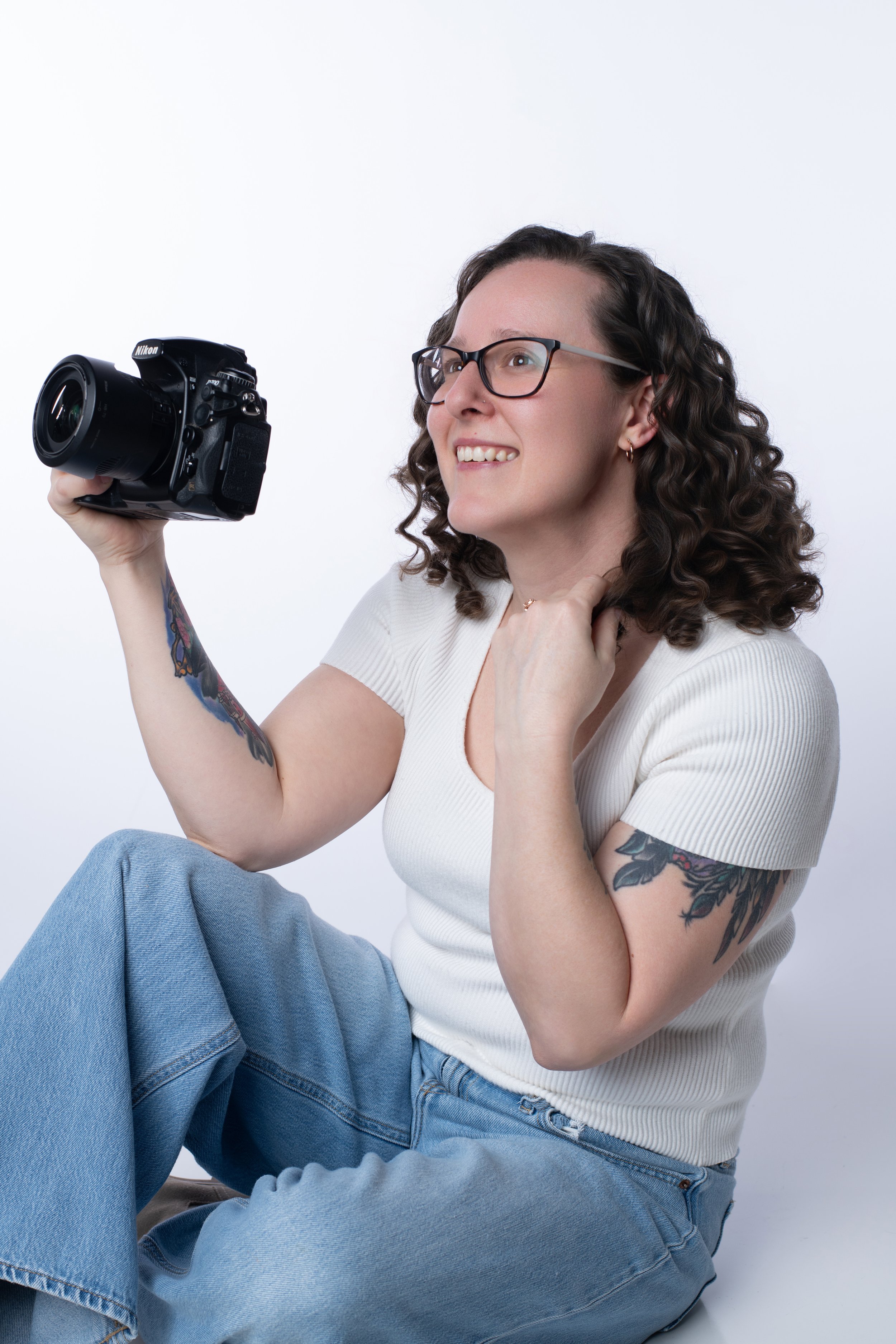 A young woman with curly brown hair, glasses, and tattoos on her arm, sitting cross-legged, holding a Nikon camera, and smiling.