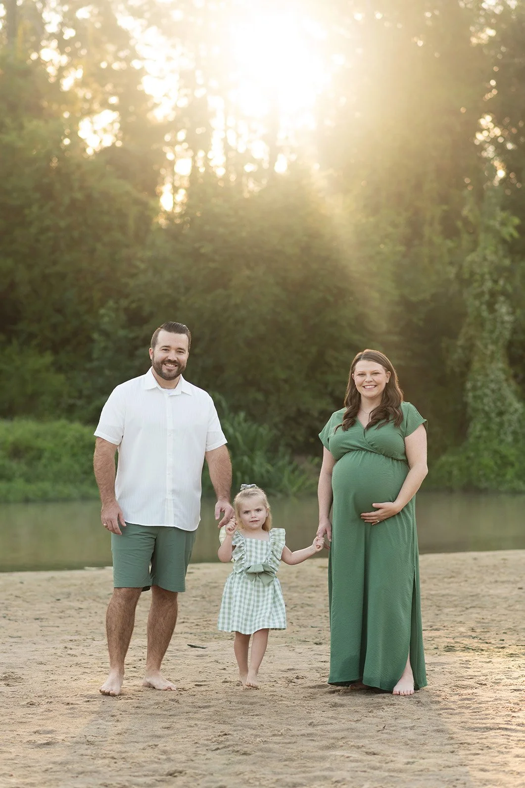 A family of three standing on a sandy beach at sunset, with a lake and green trees in the background. The pregnant woman is wearing a green dress, the man is wearing a white shirt and green shorts, and a young girl is wearing a green and white checke