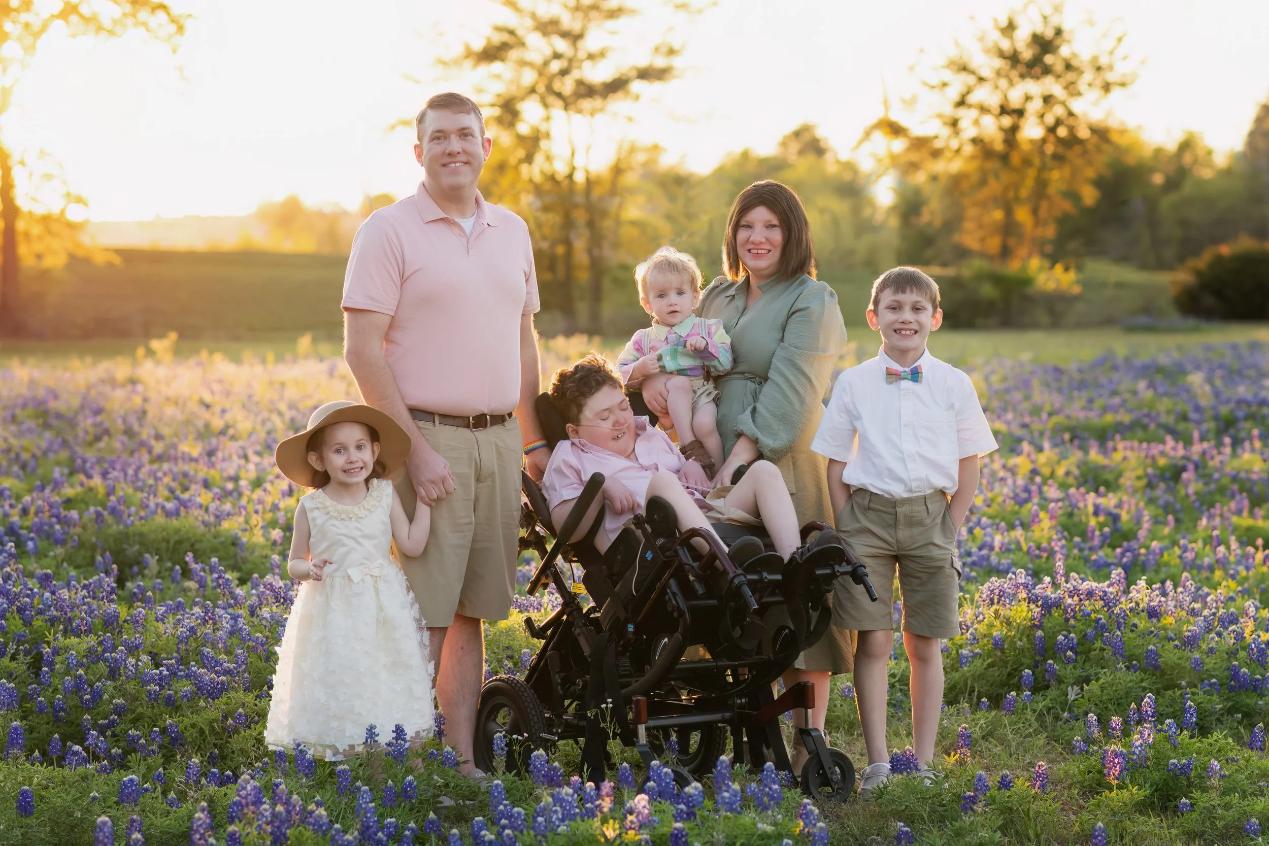 A family of seven, including children and adults, are standing and sitting in a field of purple and blue flowers during sunset. Some children are wearing formal clothing, and one adult is in a pink polo shirt. They are smiling and enjoying the outdoo