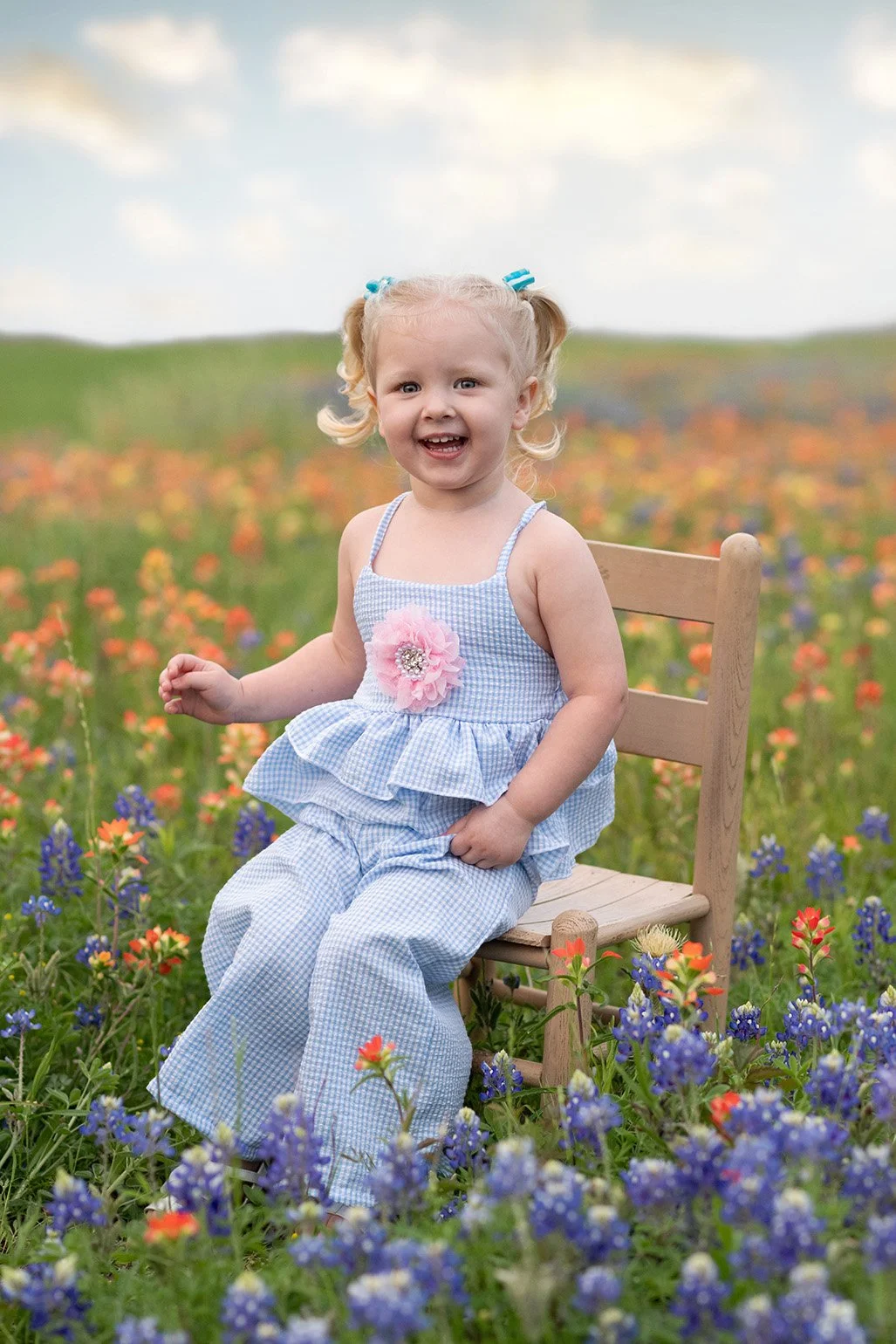 A young girl sitting on a wooden chair in a colorful flower field, smiling and wearing a blue and white gingham dress with a pink flower embellishment.