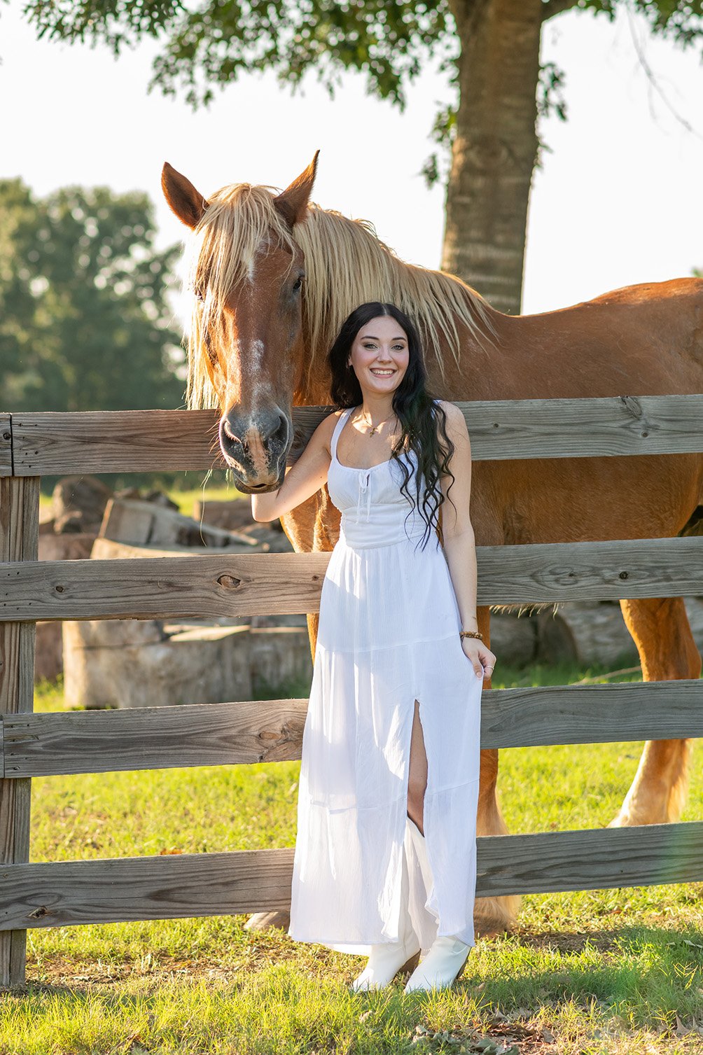 A woman with long dark hair in a white dress is standing next to a brown horse with a blonde mane, holding its bridle in her right hand, outdoors near a wooden fence with green grass and trees in the background.