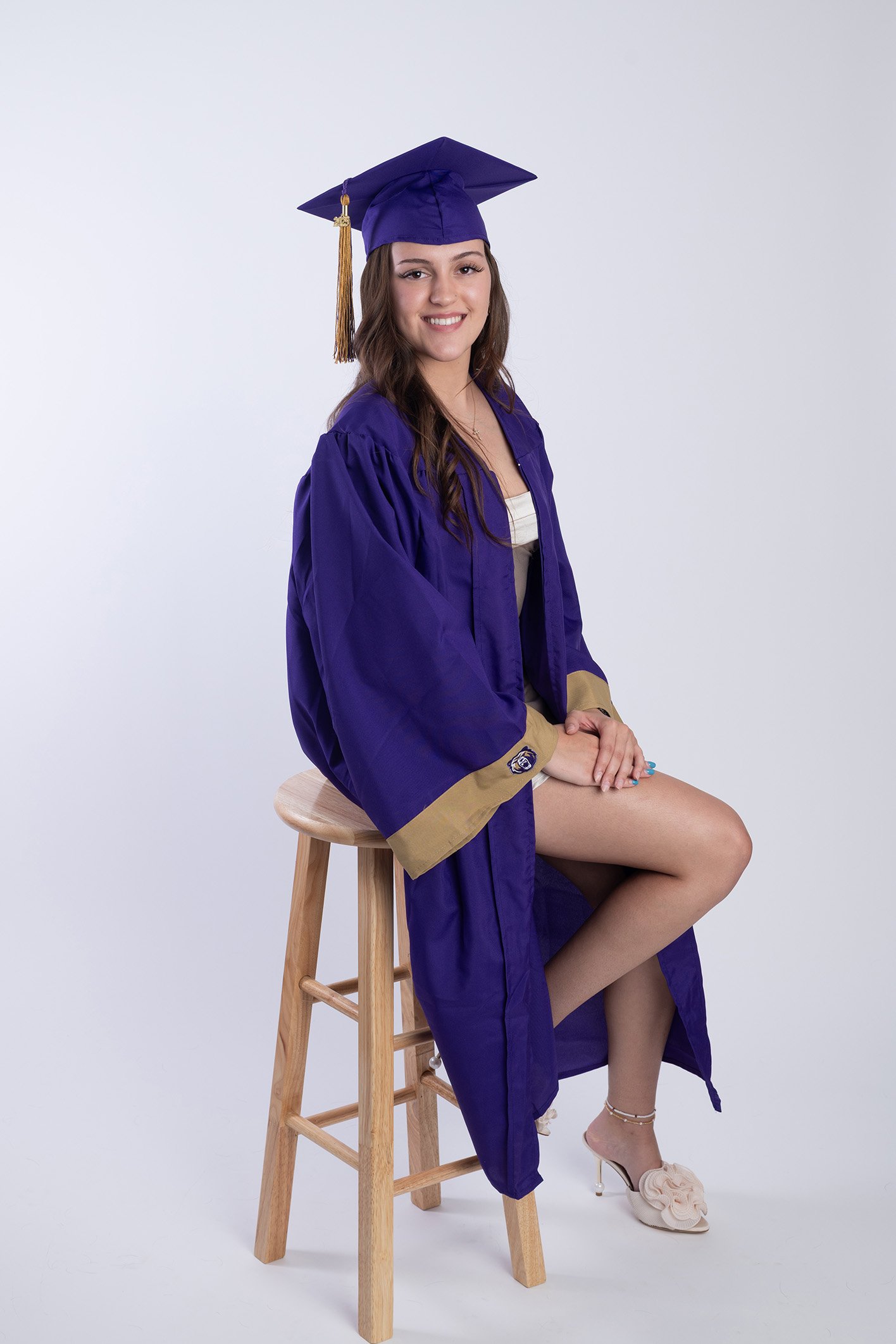 Young woman in a graduation cap and gown sitting on a wooden stool with her right leg crossed over her left, smiling. She is wearing beige shoes with decorative bows and white socks. The background is plain white.