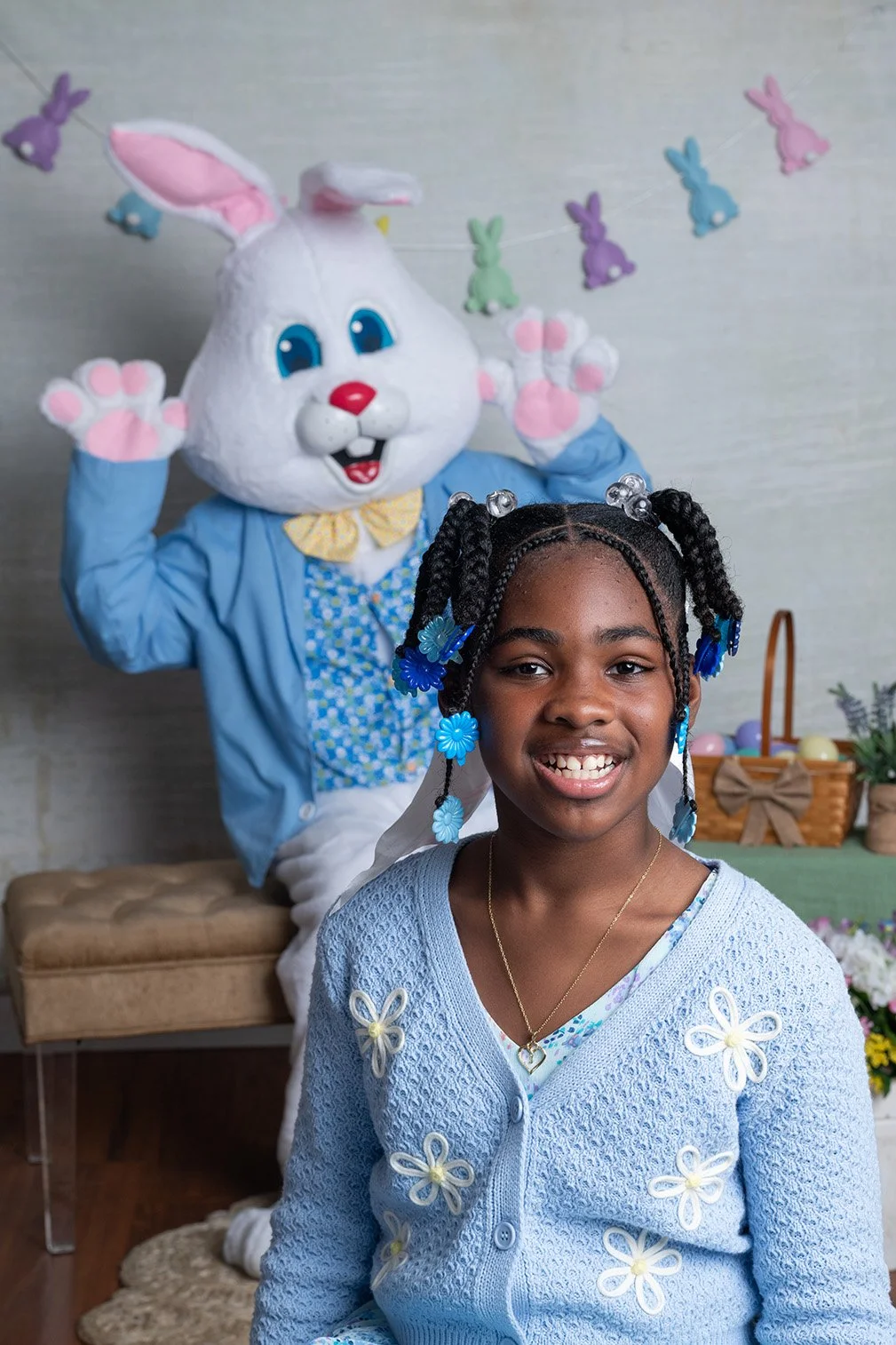 A young girl with braided hair and colorful hair accessories smiling in front of an Easter-themed display, with a person in a white bunny costume dressed in a blue jacket and floral shirt behind her.