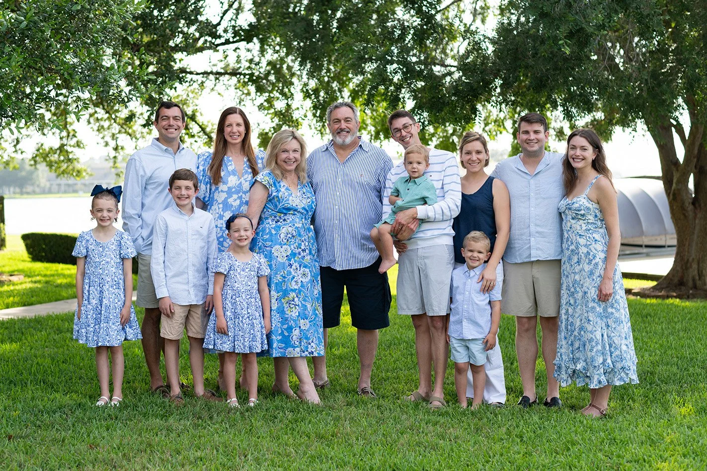 Family gathering outdoors under a large tree, with adults and children smiling and dressed in blue and white, standing on green grass.