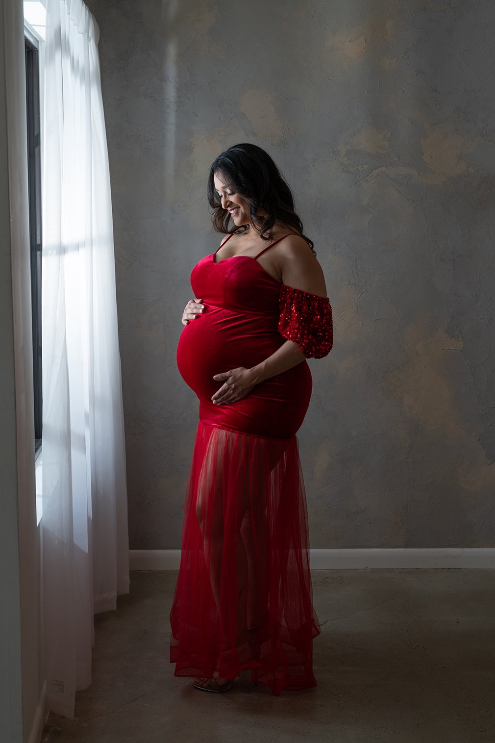 A pregnant woman in a red dress standing near a window with white curtains, smiling and gently touching her belly.