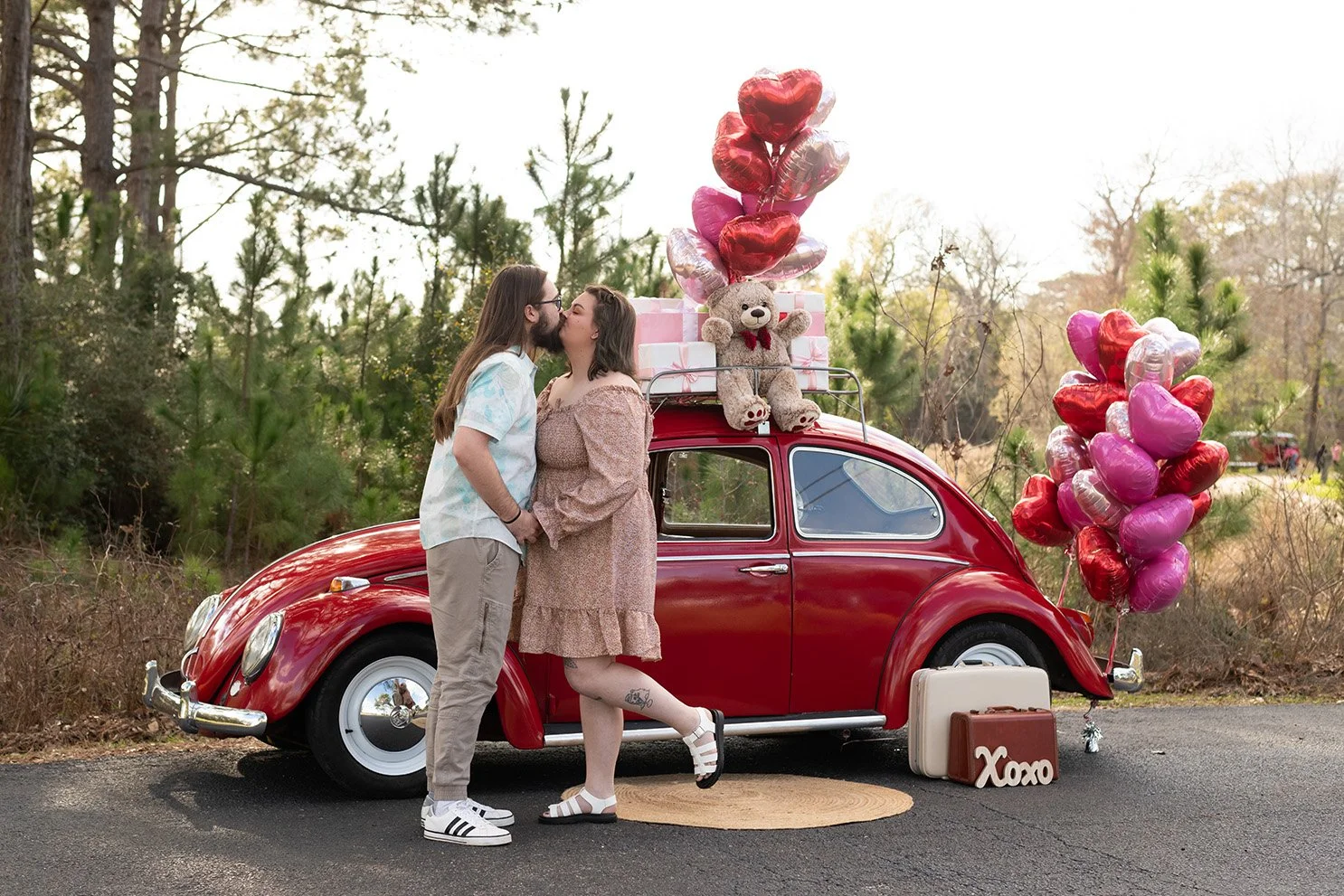 A couple kissing in front of a red vintage car decorated with pink and red heart-shaped balloons, teddy bear plush, and gift boxes, outdoors with trees in the background.