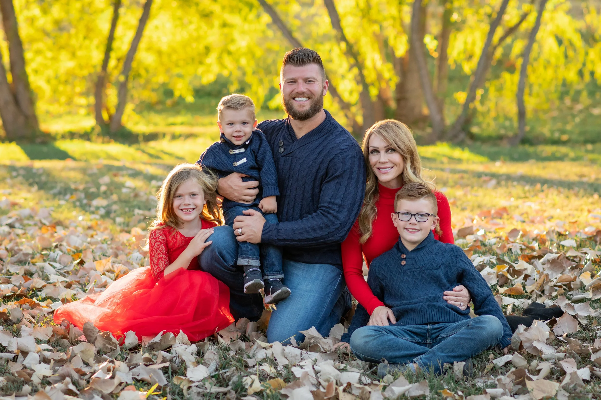 A family of six posing outdoors in a park during fall, surrounded by fallen leaves and yellow trees, with sunlight filtering through the leaves.