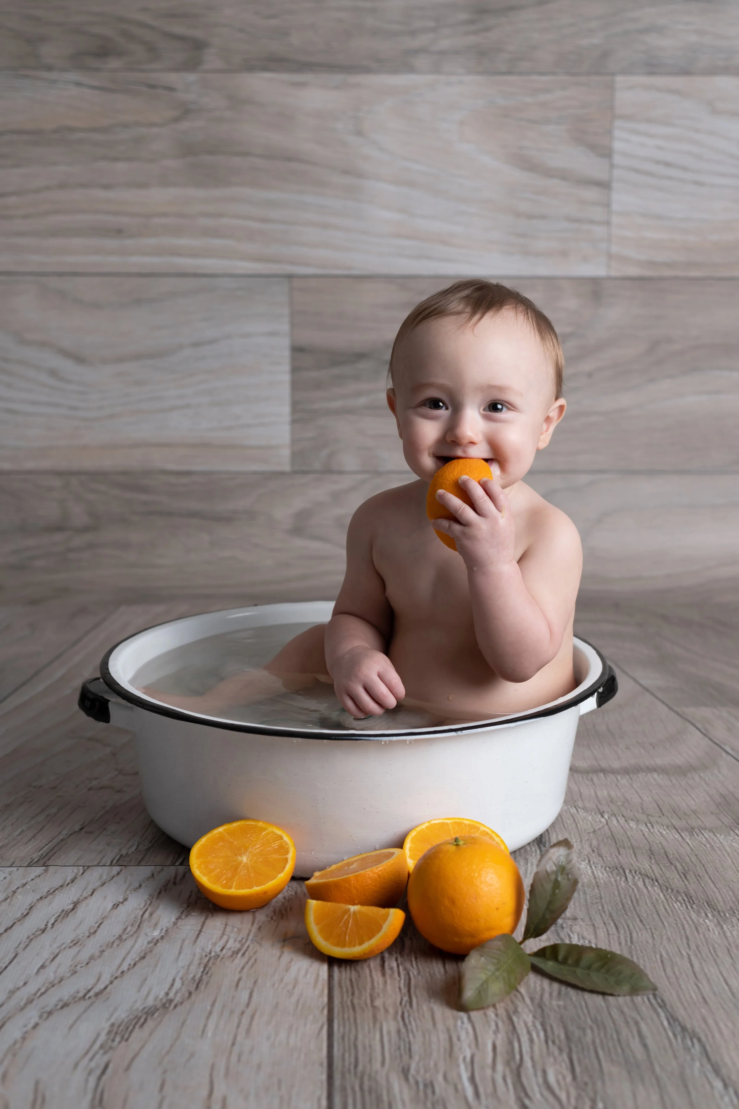 A smiling baby sitting in a white basin filled with water, holding a halved orange, with more halved oranges and green leaves on a wooden floor.