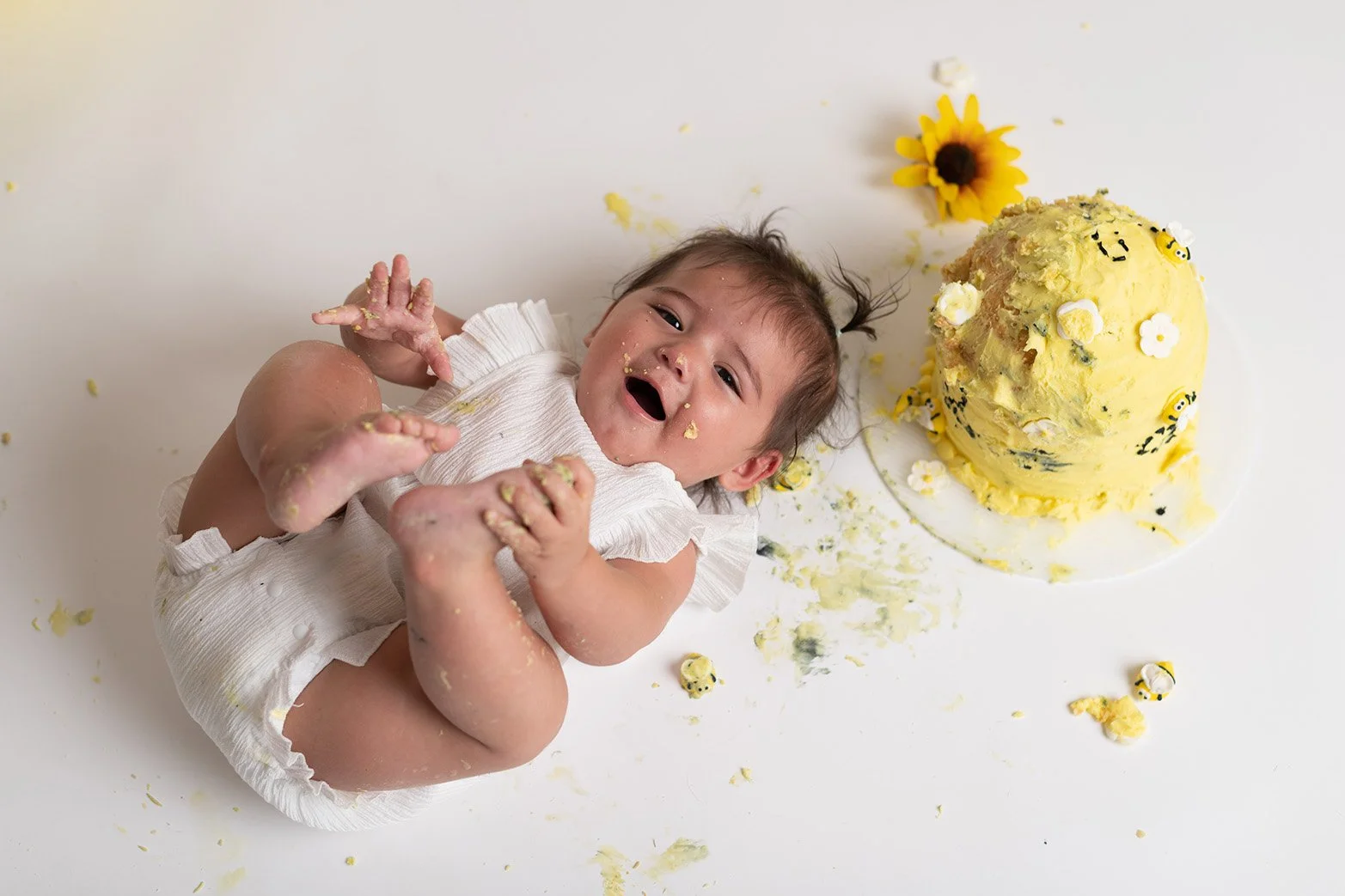 A baby lies on a white surface covered in yellow cake and frosting, smiling with cake on their face and hands, next to a partially eaten yellow cake decorated with small white and black bee-themed decorations and sunflower on the side.
