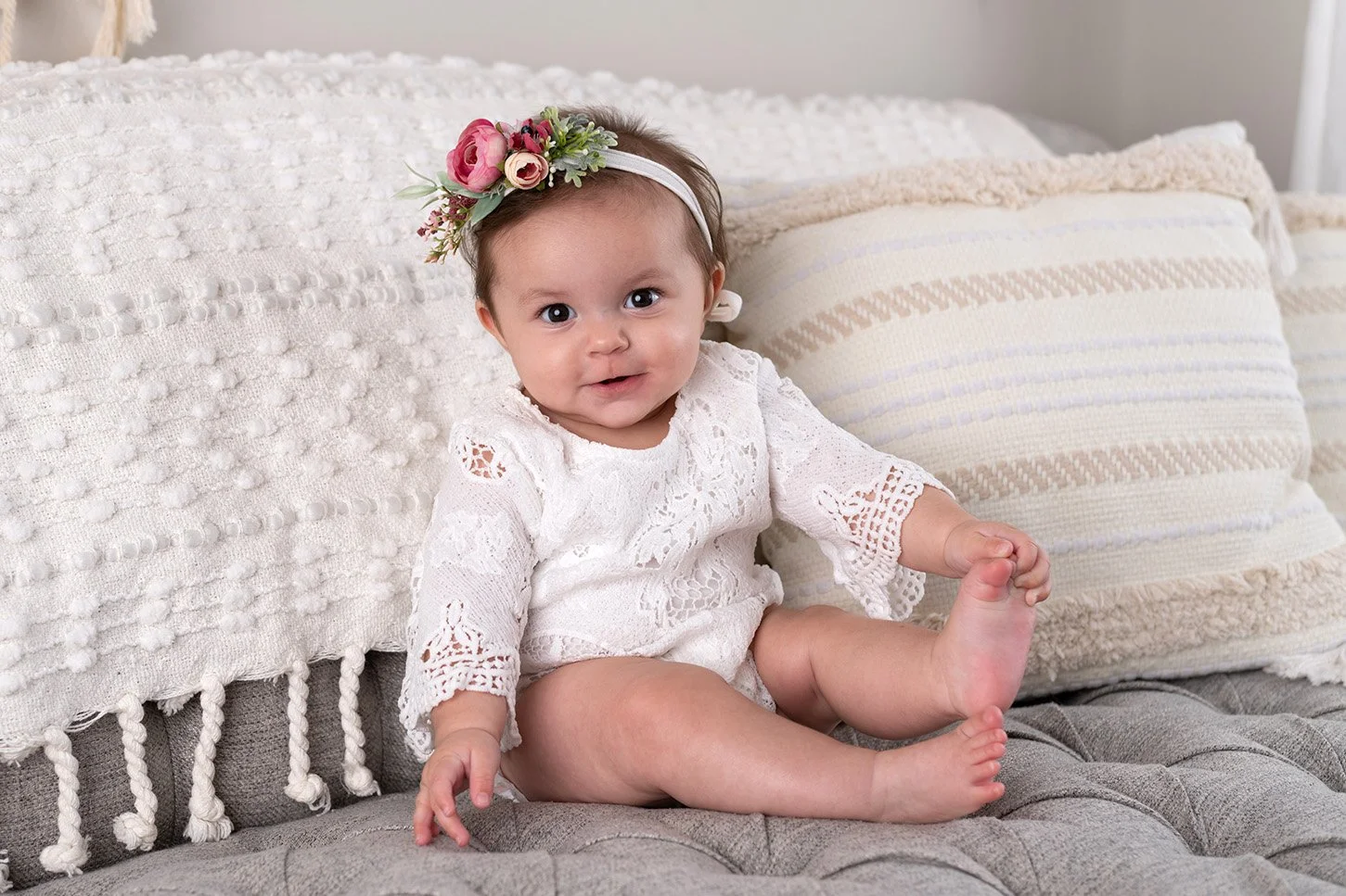 Adorable baby girl sitting on a gray upholstered bench, wearing a white lace dress and a floral headband, surrounded by cream-colored pillows.