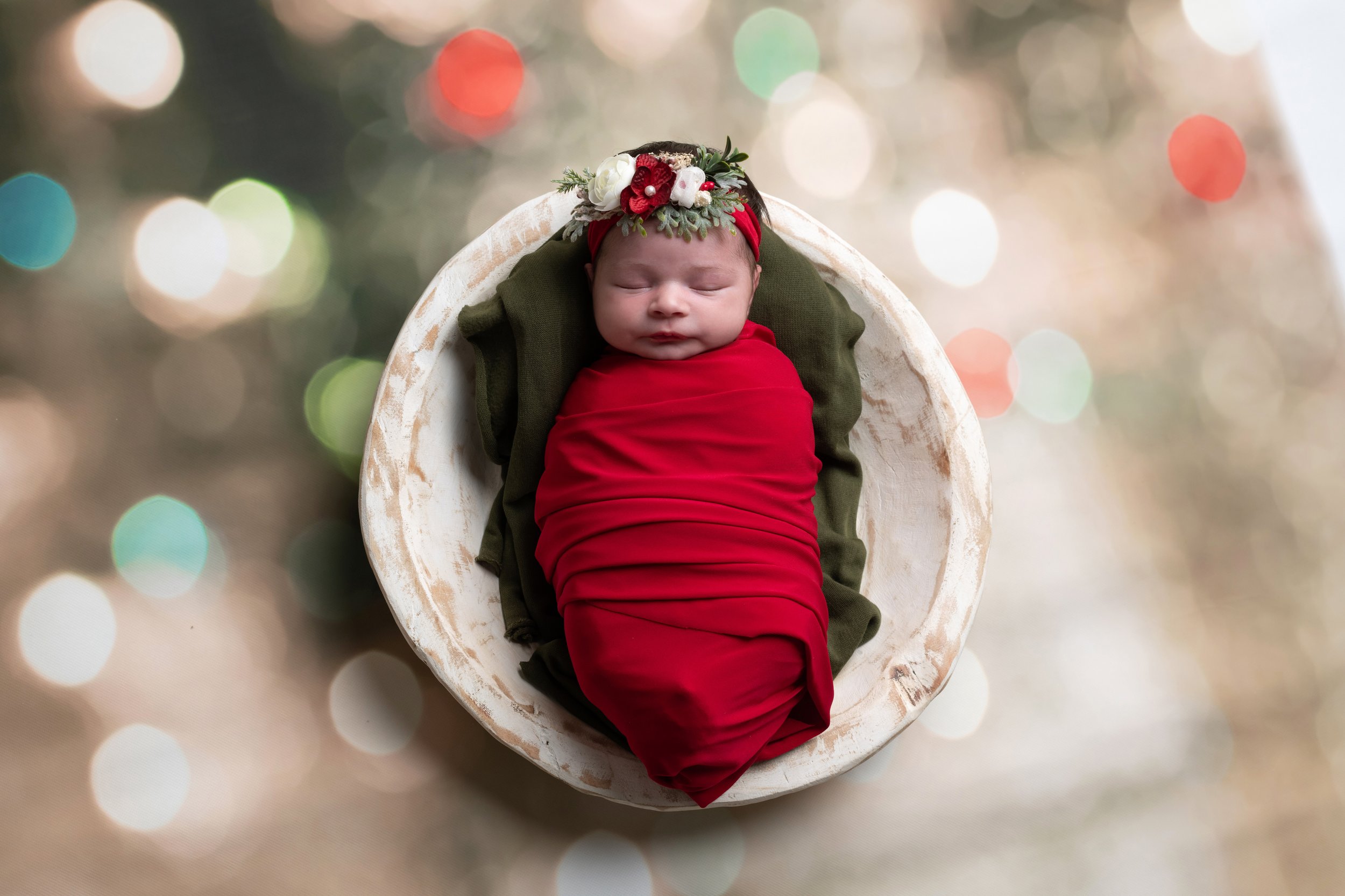 A sleeping infant dressed in red and green, lying in a white oval-shaped bowl, wearing a floral headband with red and white flowers and greenery, against a bokeh background of multicolored lights.