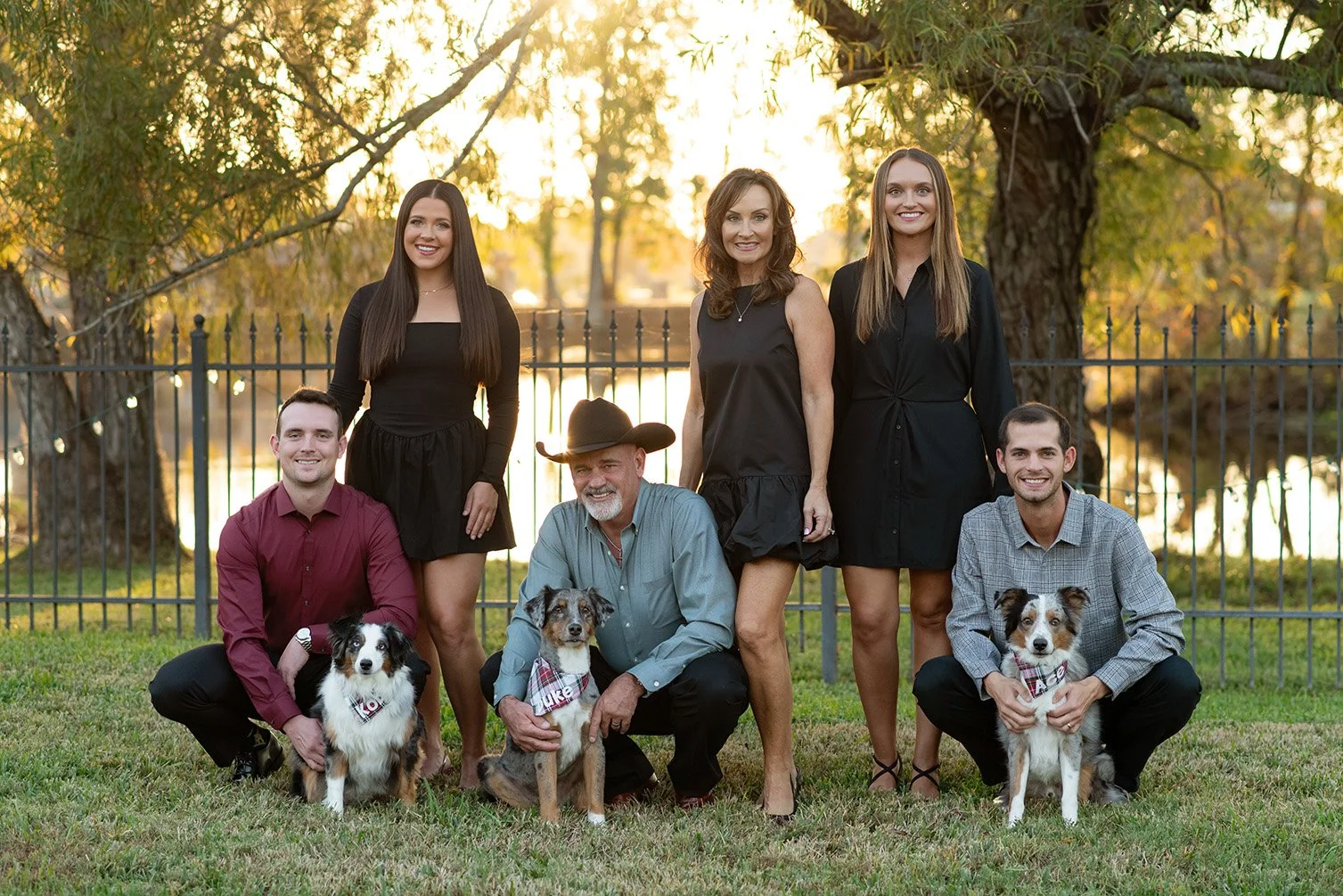 A family photo taken outdoors during sunset, featuring three women, three men, and three dogs, with a black fence and trees in the background.