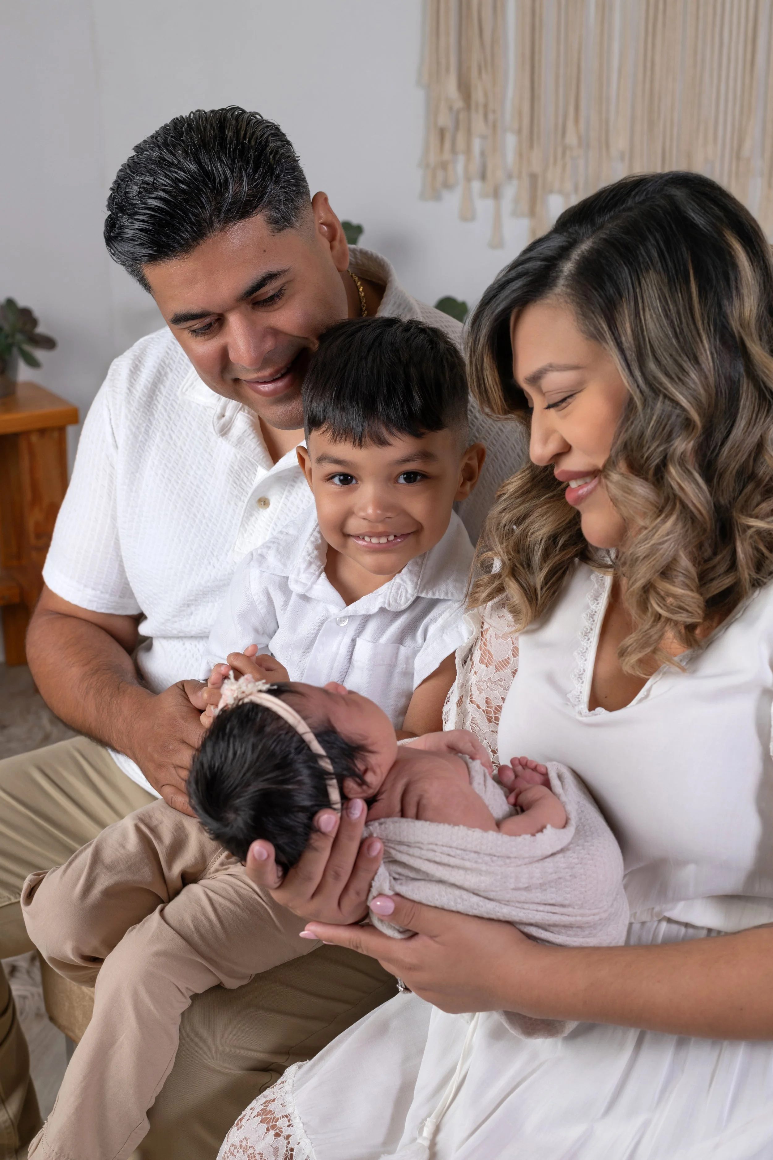 A family of four with two parents and two children, gathered together indoors, smiling and looking lovingly at a newborn baby.