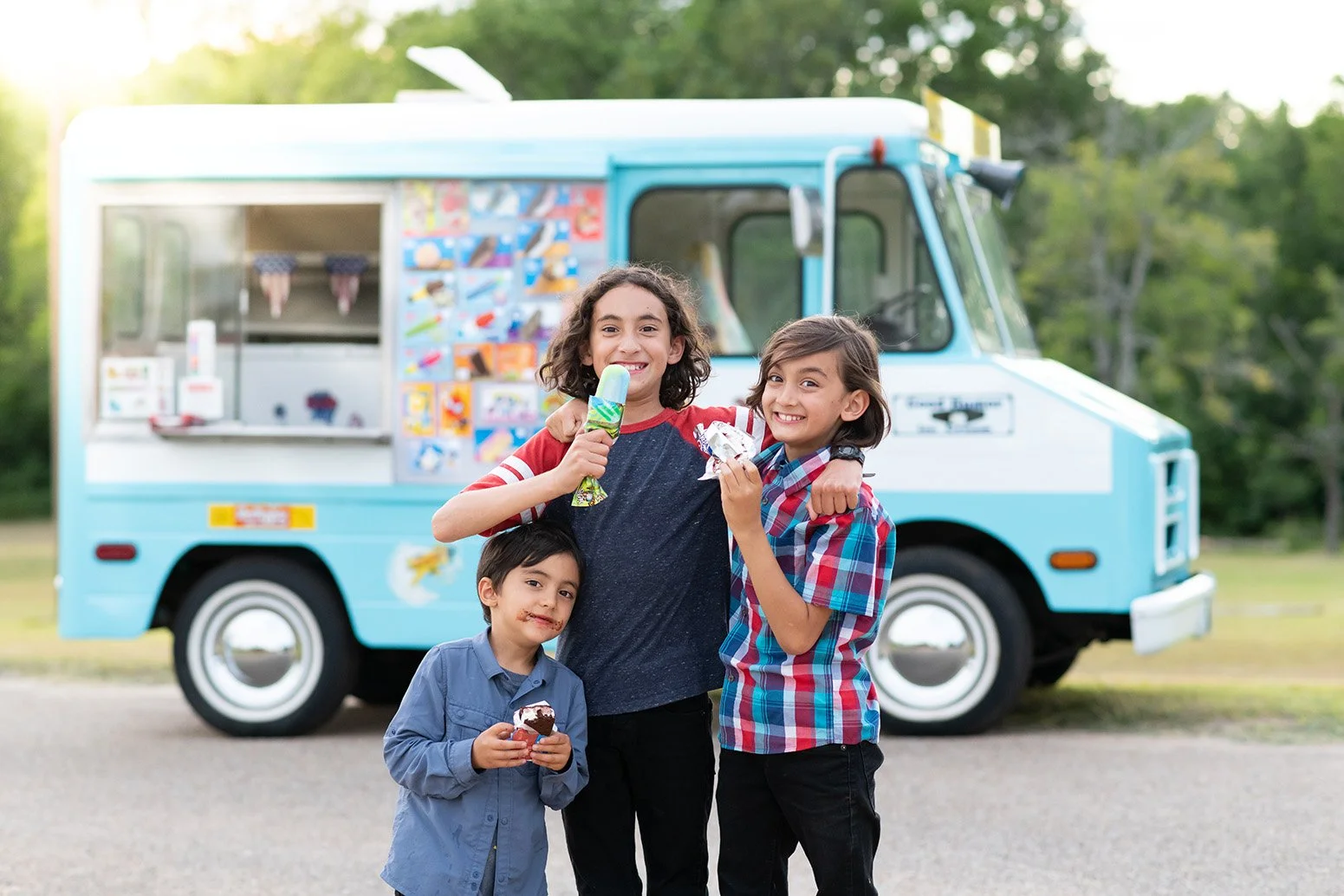 Three children standing in front of an ice cream truck, smiling and holding ice cream cones and a snack, outdoors with trees in the background.