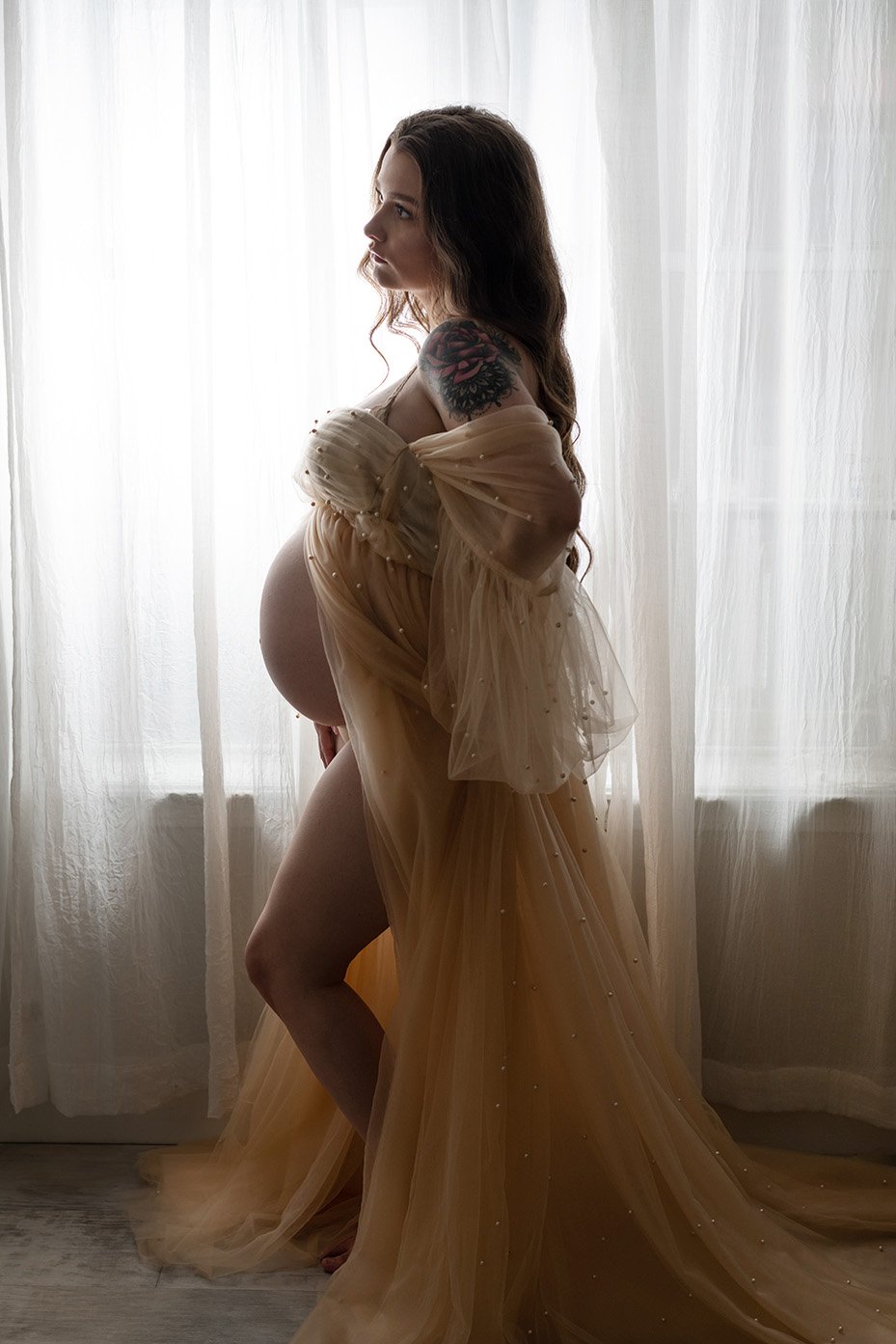 Pregnant woman standing by a backlit window wearing a flowing beige gown with sheer fabric and pearl details, displaying a large colorful floral tattoo on her shoulder.