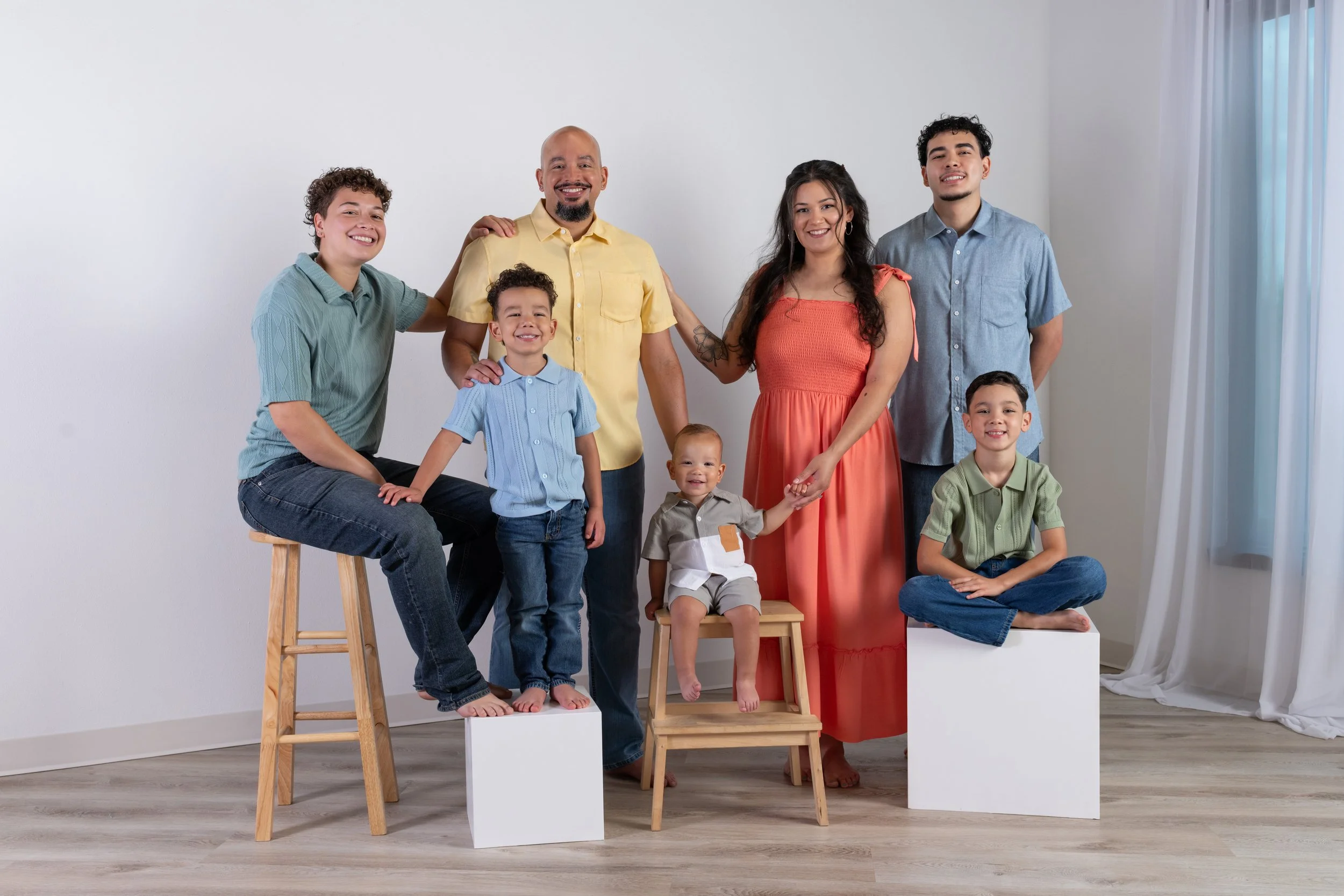 A diverse family group poses together indoors, with some standing on benches and others sitting or kneeling, smiling at the camera, against a plain white wall with natural light from a window.
