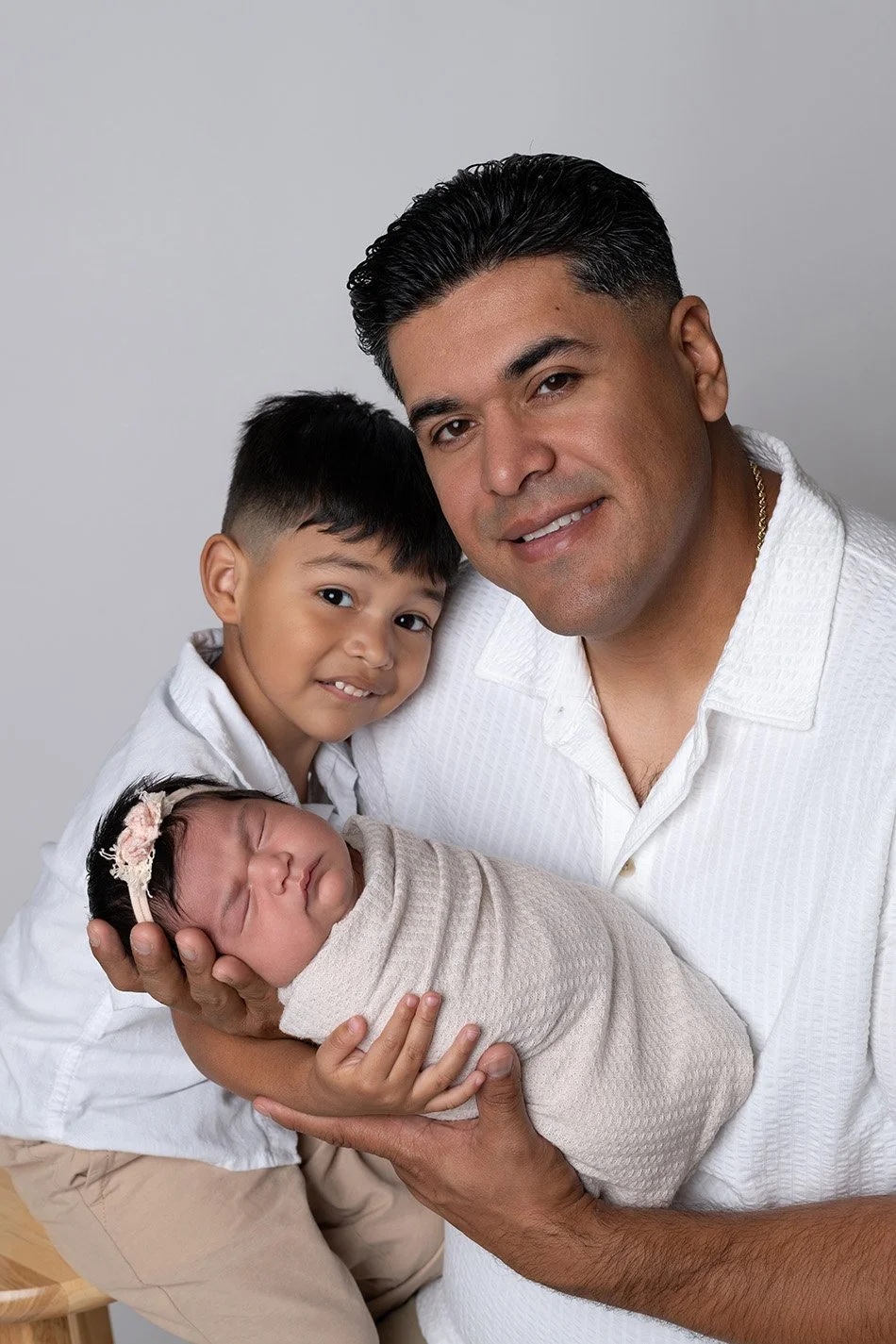 A man holding a newborn baby wrapped in a beige blanket, with a young boy leaning close, all smiling at the camera against a neutral background.