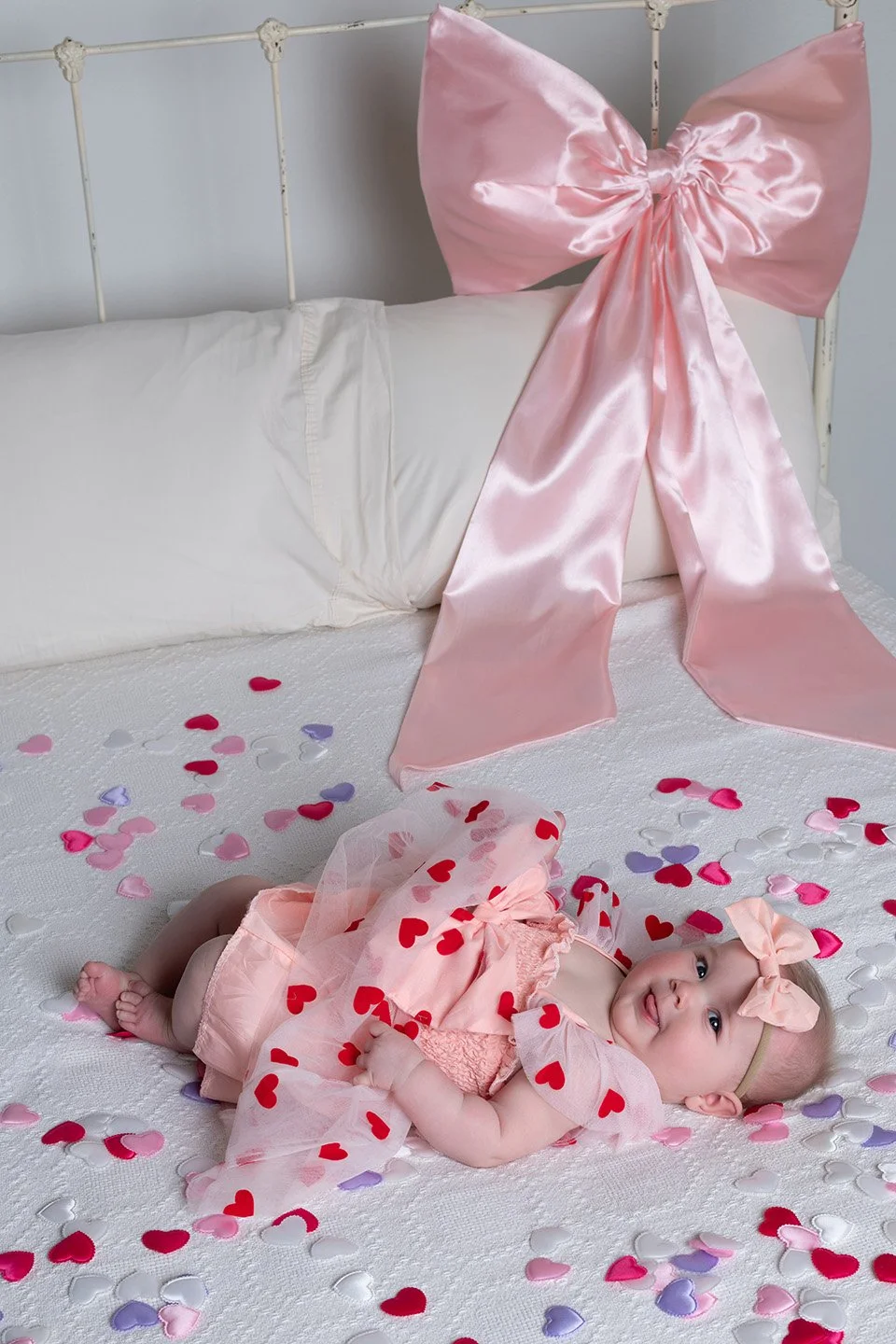 A baby girl lying on a white bed covered with pink, purple, red, and white heart-shaped confetti. She is dressed in a pink dress with matching headband with a bow, and is surrounded by scattered hearts.