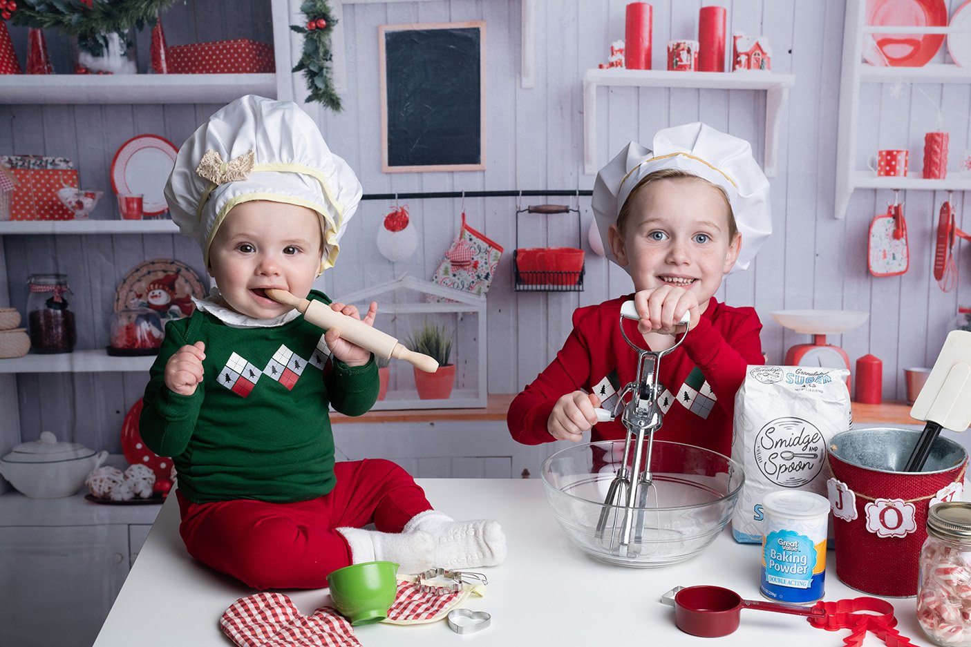Two children dressed as chefs, wearing festive sweaters and chef hats, baking together in a holiday-themed kitchen with red and white decorations.