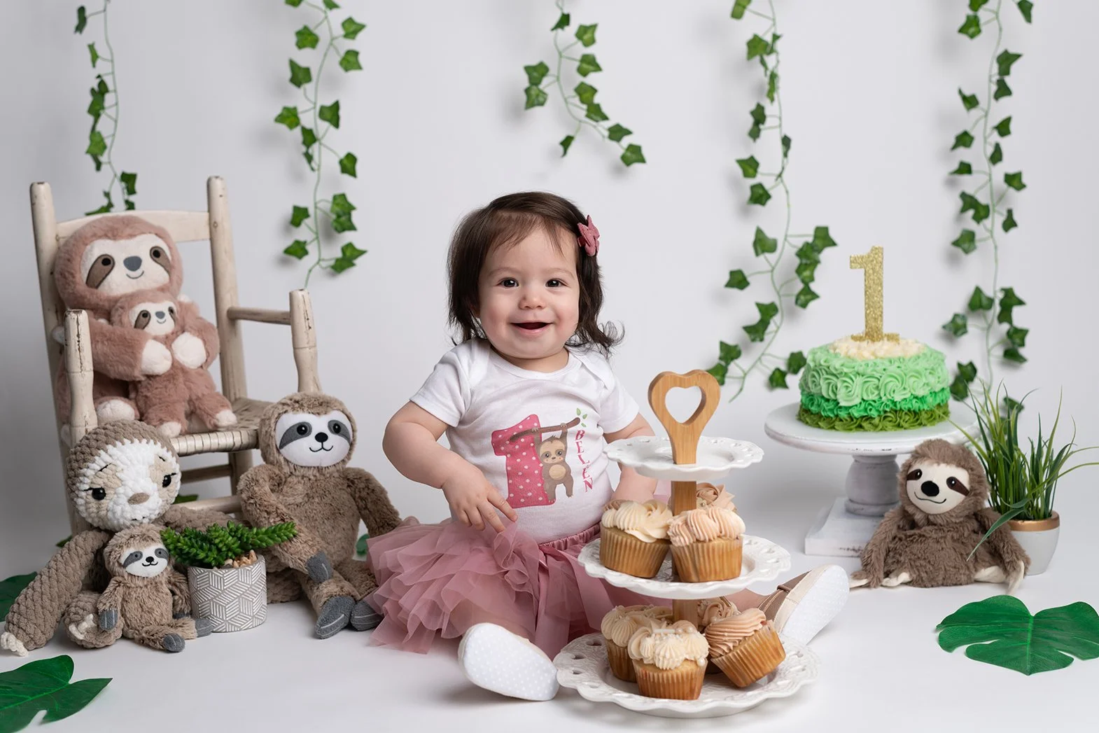 A young girl celebrating her first birthday with a cake, cupcakes, and plush sloth and lion toys, surrounded by green leaves and vines.