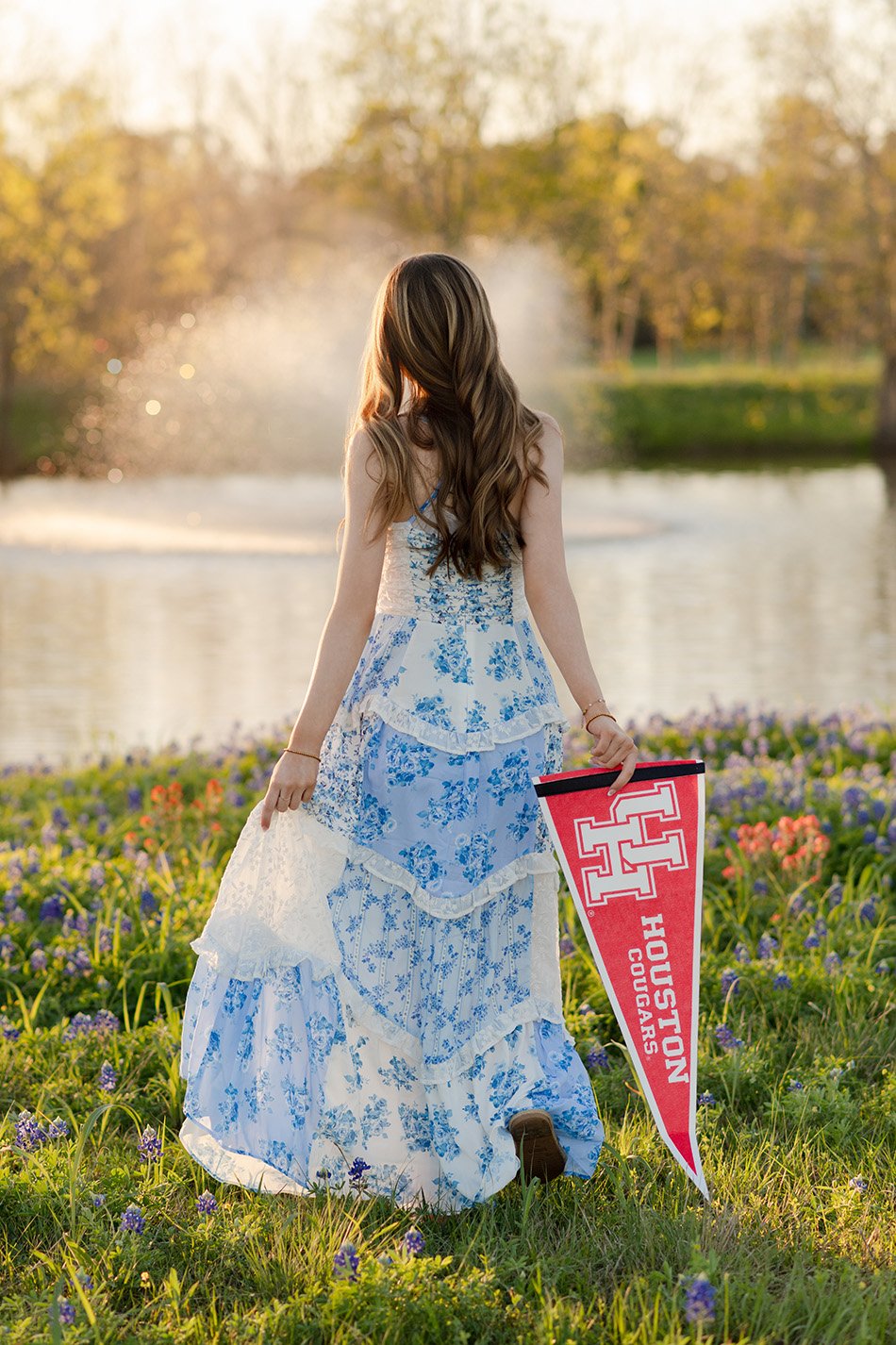 A woman with long brown hair in a floral blue and white dress, standing in a field of purple and yellow flowers beside a body of water, holding a red Houston Cougars pennant, with a fountain and trees in the background during sunset.