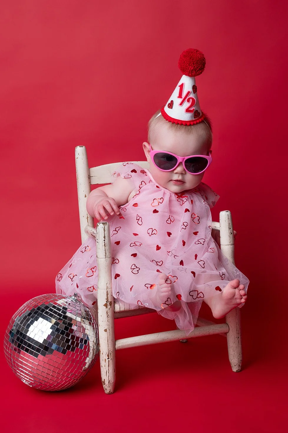 A young child wearing pink sunglasses, a pink dress with red and pink hearts, and a party hat with '1/2' and red hearts, sitting on a white wooden chair against a red background. A disco ball is on the floor beside her.