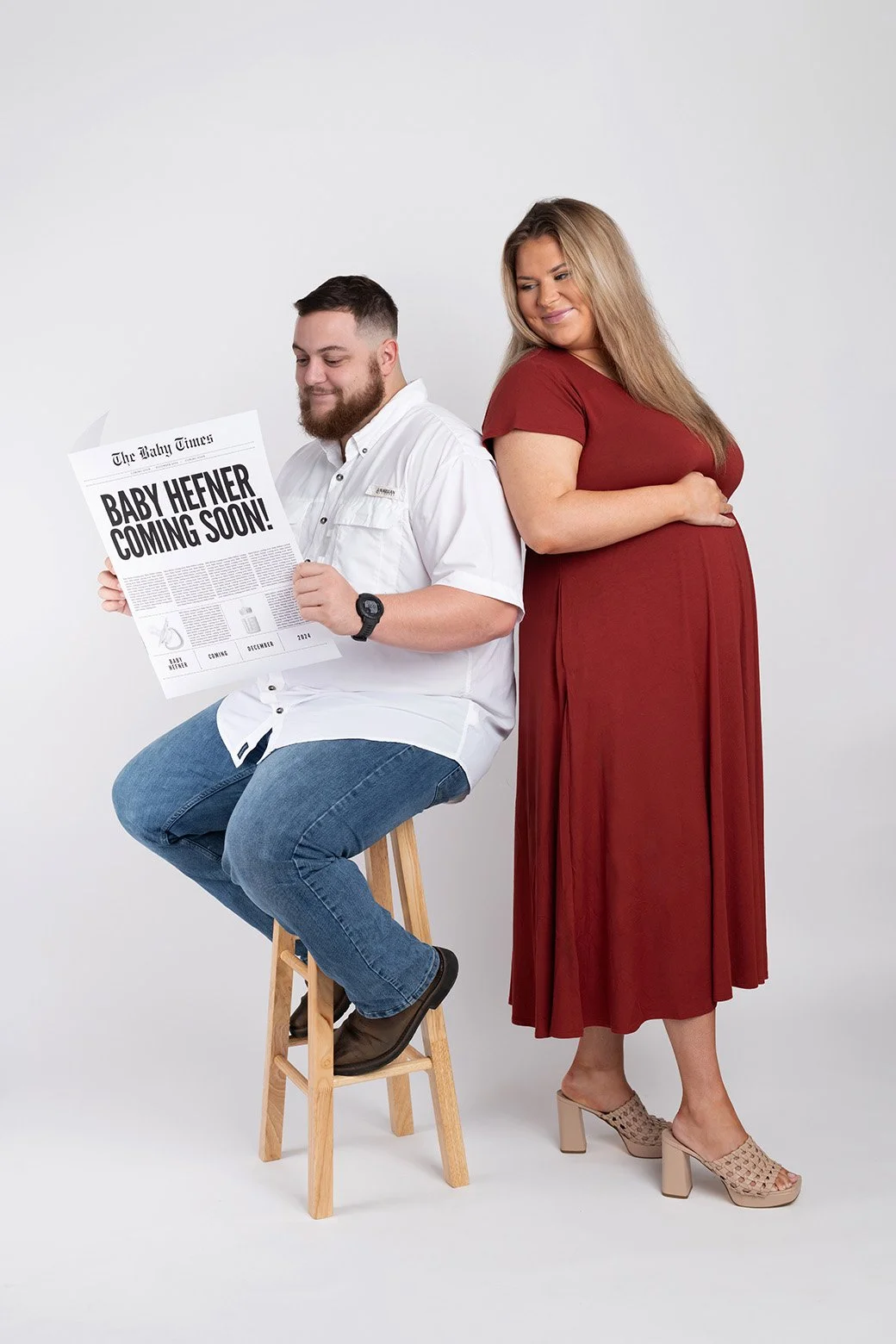 A man sitting on a stool holding a newspaper that reads 'Baby Heifer Coming Soon!' while a pregnant woman in a red dress stands next to him, smiling with her hand on her belly against a plain white background.