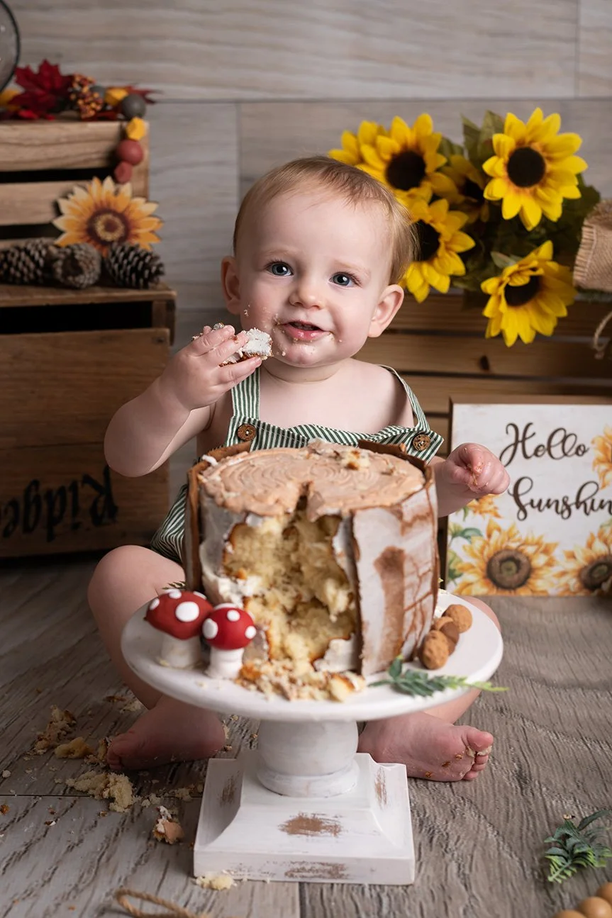 A young child with short hair and blue eyes sitting in front of a partially eaten vanilla cake with cream frosting, holding a piece of cake in his hand. The child is smiling and has crumbs and frosting on his face. The cake is decorated with red and 