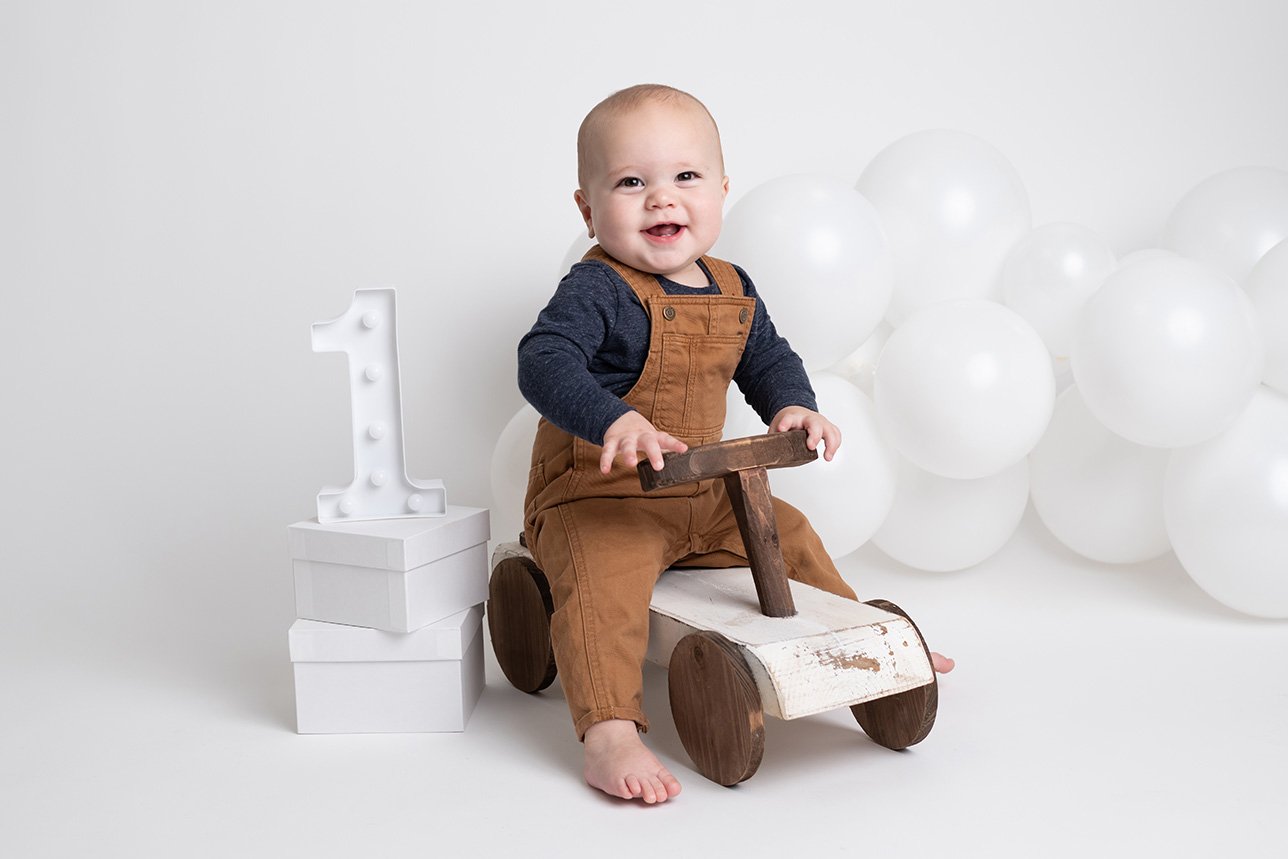 A smiling baby boy riding a wooden toy car with white balloons and a white number one decoration in the background.