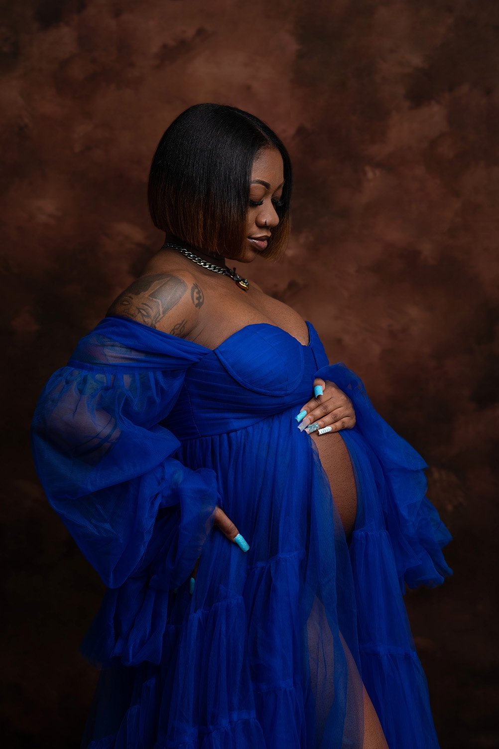 A pregnant woman with tattoos on her left shoulder and arm, wearing a blue dress with sheer fabric, standing against a brown mottled background.
