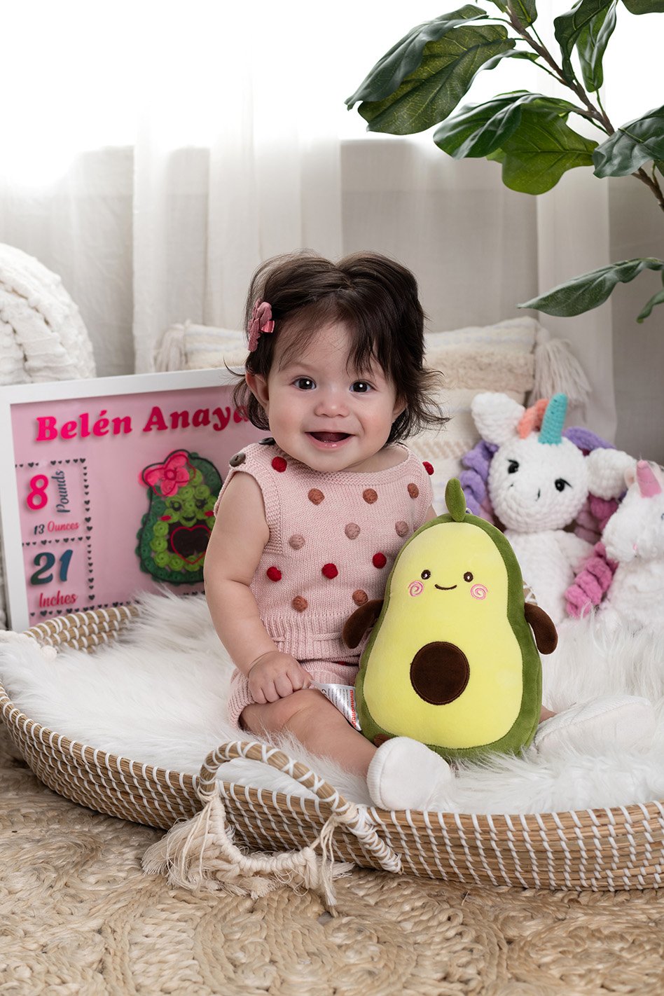 A smiling baby girl sitting in a wicker basket with plush toys, including an avocado-shaped stuffed animal, in a cozy, decorated room with a plant and a pink milestone display in the background.