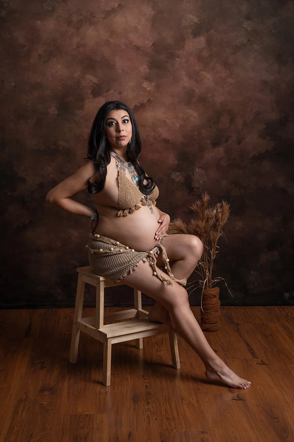 A pregnant woman with long dark hair sits on a wooden stool in a photography studio. She wears a handmade crochet top and skirt with beaded accessories and poses with one hand on her belly and the other behind her back. The background is a textured b