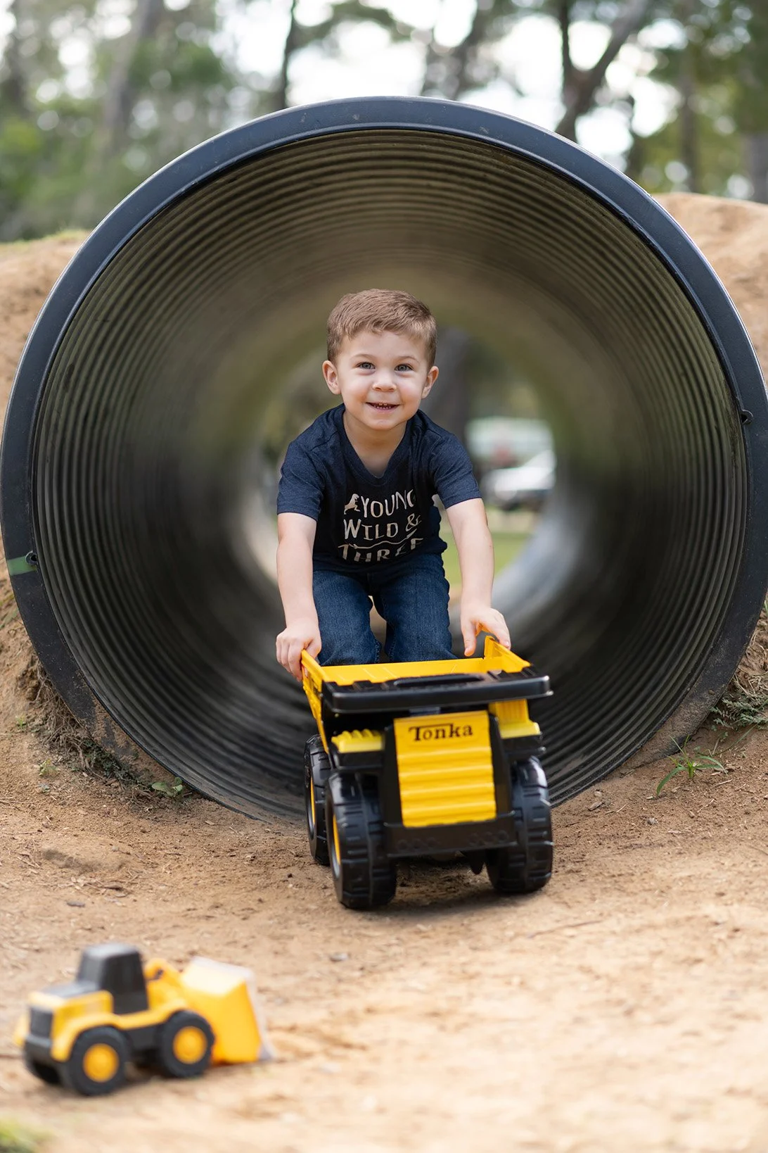 A young boy playing inside a large metal pipe at a park, pushing a yellow toy dump truck with the name 'Tonka' on it. He is smiling and wearing a dark t-shirt and jeans, with toy construction vehicles on the ground nearby.