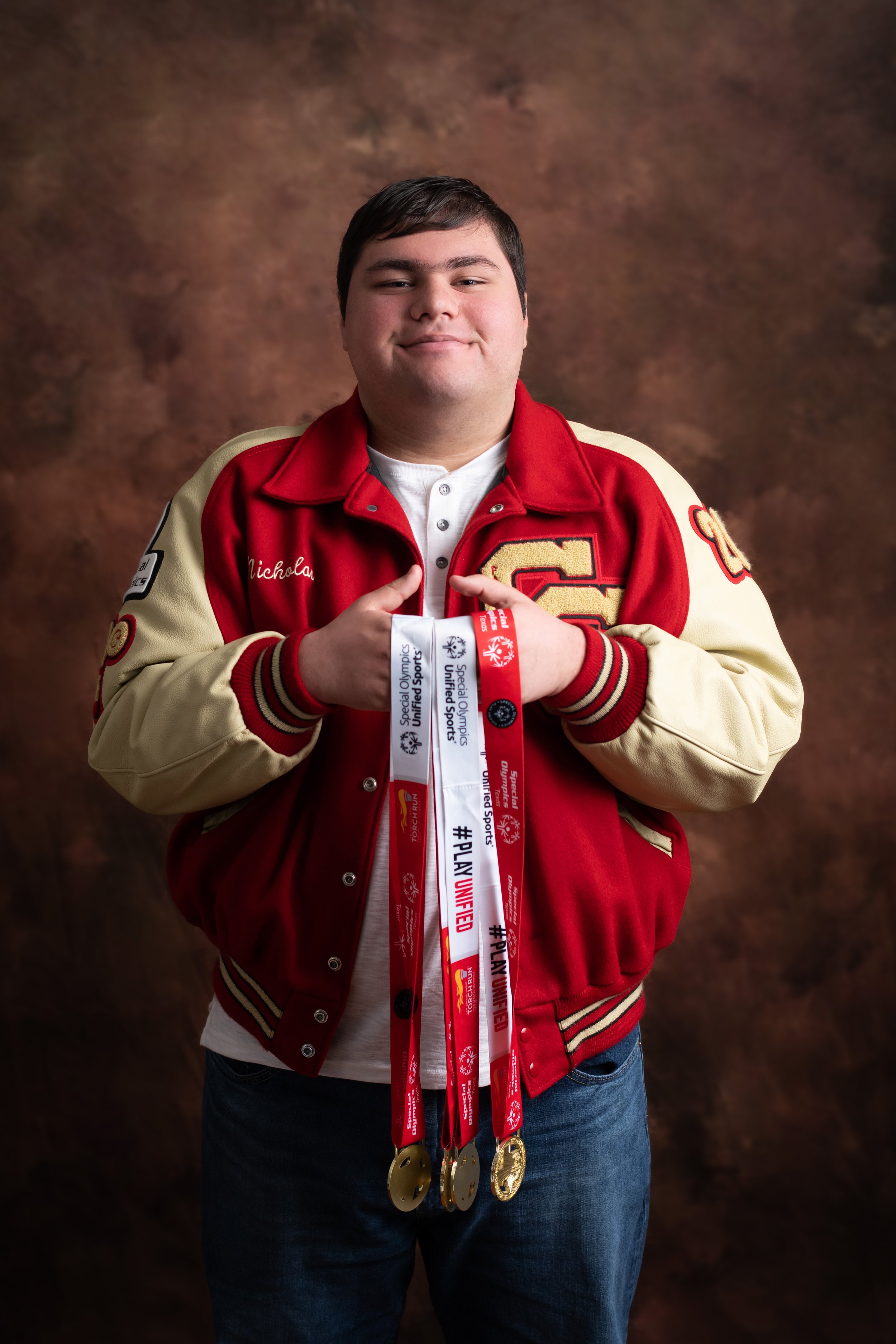 A young man with short dark hair is smiling and holding up medals and ribbons, wearing a red and cream varsity jacket. The background is brown and textured.