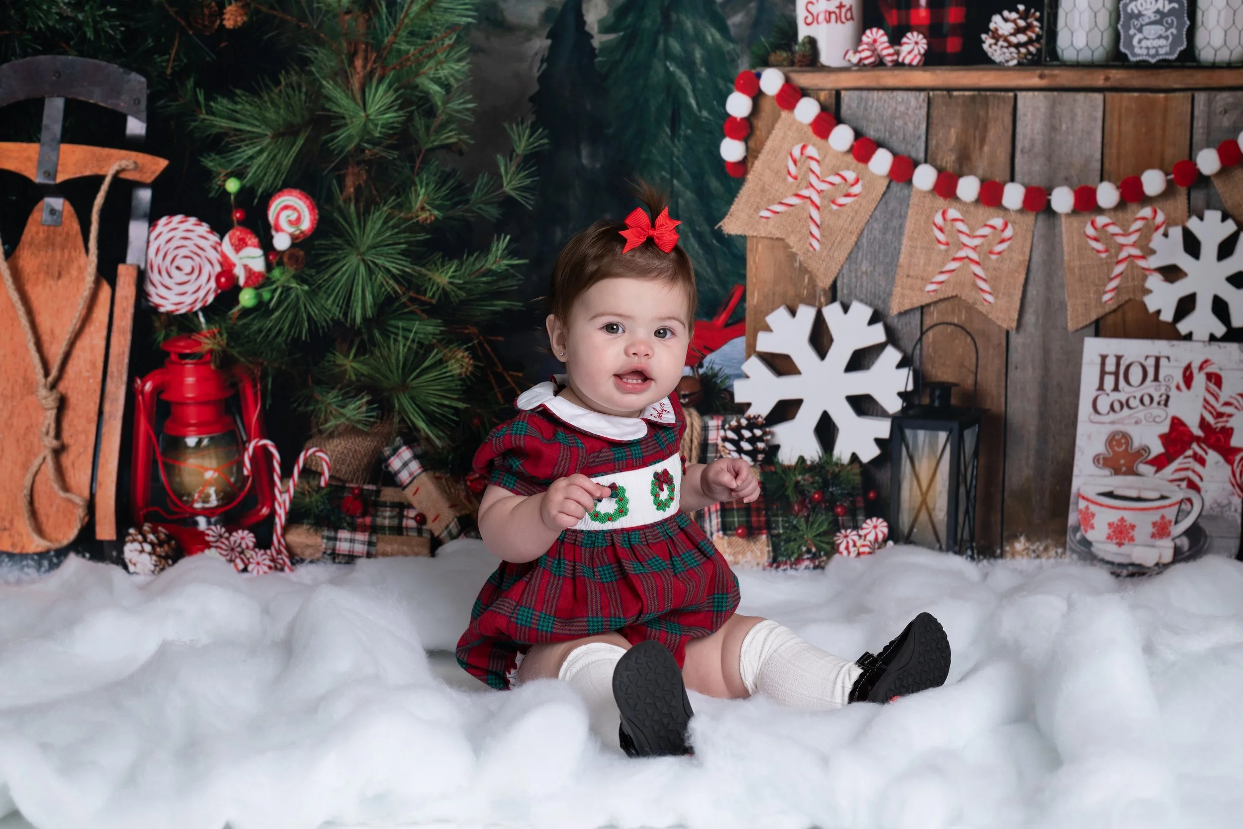 A young child wearing a red and green Christmas dress with holiday decorations, including a decorated Christmas tree and festive ornaments, sitting on faux snow at a holiday scene.