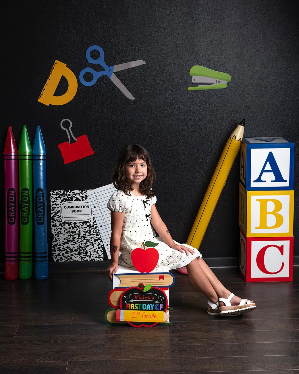 A young girl sitting on the floor in a classroom-themed setting with large foam school supplies and colorful pencils. There are a chalkboard wall with cutouts of scissors, a stapler, and a stapler, and a stack of alphabet blocks spelling ABC. She is 