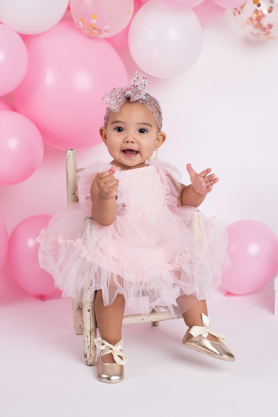 A baby girl sitting on a small wooden chair, wearing a pink tulle dress, shiny gold shoes with bows, and a sparkly pink headband with a bow. She is surrounded by pink and white balloons and looks happy.