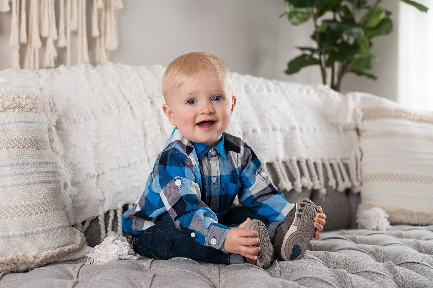 A smiling young boy with blonde hair, wearing a blue and gray plaid shirt, sitting on a plush, tufted gray cushion on a sofa with decorative pillows, in a cozy living room with a plant in the background.