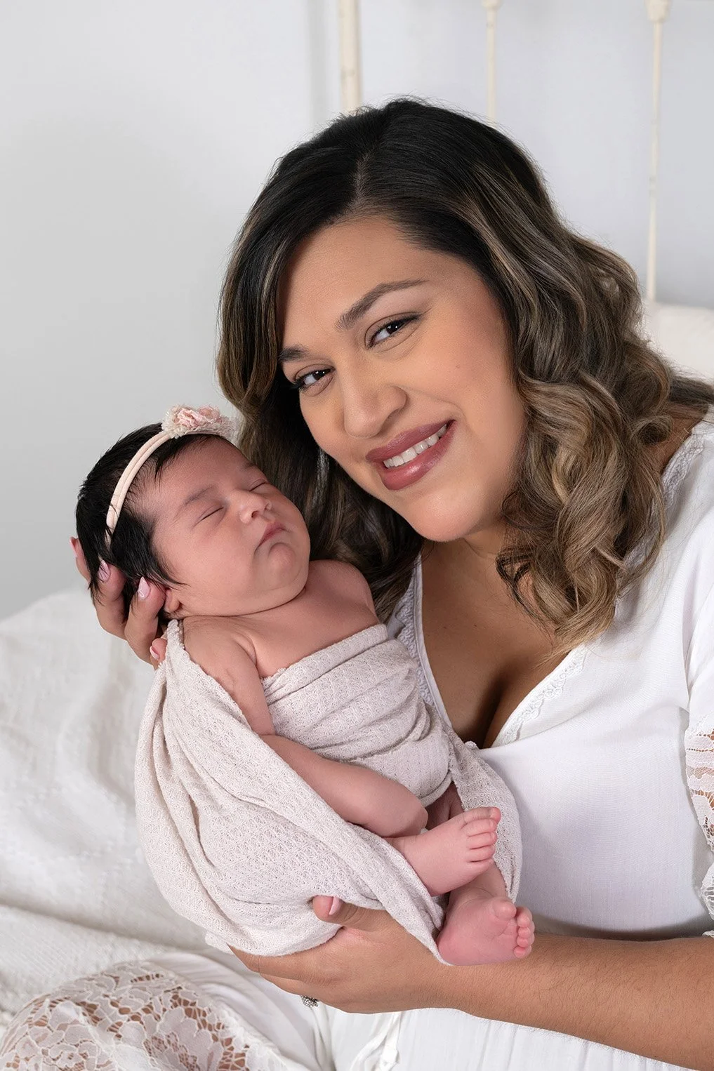 A woman with wavy brown hair and wearing a white top is holding a sleeping newborn baby wrapped in a beige blanket. The woman is smiling gently at the camera.