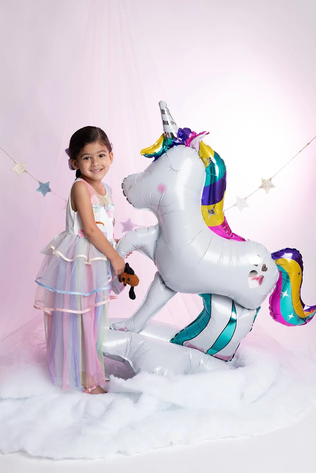 A young girl in a pastel rainbow-colored dress standing next to a large inflatable unicorn with rainbow-colored mane and tail, on a fluffy white surface with pink and white stars in the background.