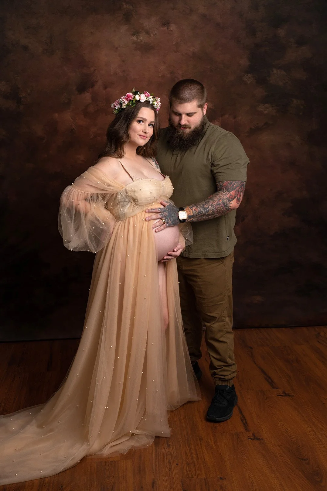 A pregnant woman in a flowing beige gown with pearl embellishments, wearing a flower crown, stands beside a man with a beard, tattoos, and a short haircut, wearing a green shirt and brown pants, both holding her belly against a brown textured backdro