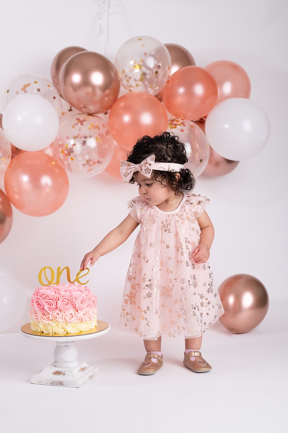 A young girl standing in front of a backdrop of pink, white, and metallic balloons, wearing a floral dress and a bow headband, reaching for a cake with a gold "one" topper.