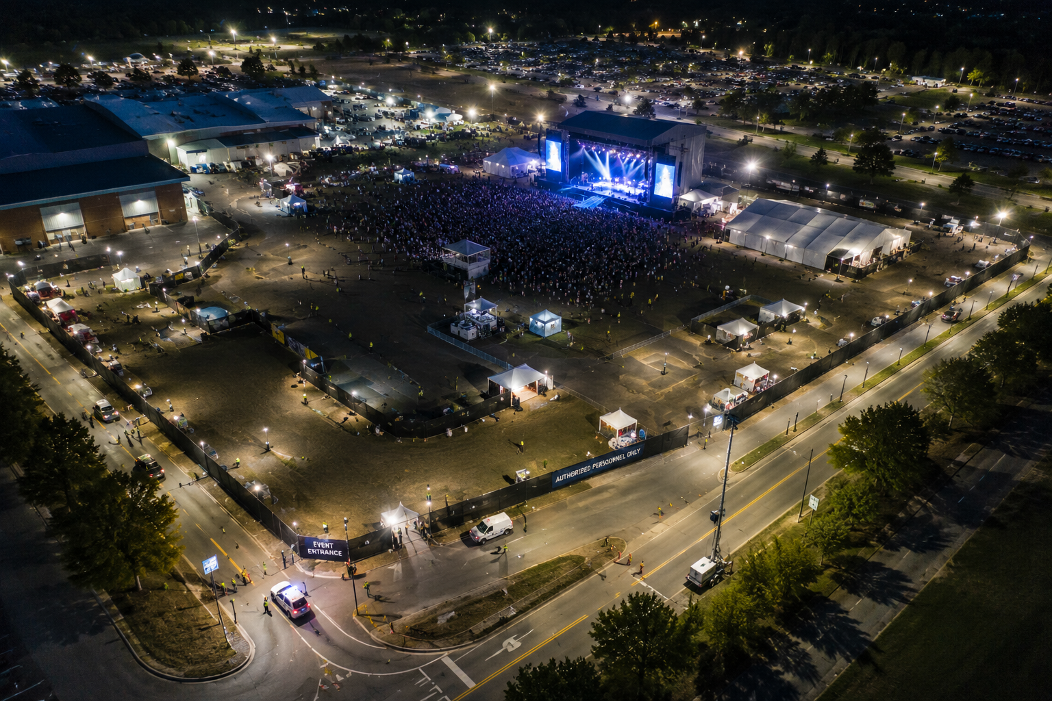 Aerial nighttime view of an outdoor concert with a large stage, bright lights, a crowd of attendees, surrounding tents, and parked cars.