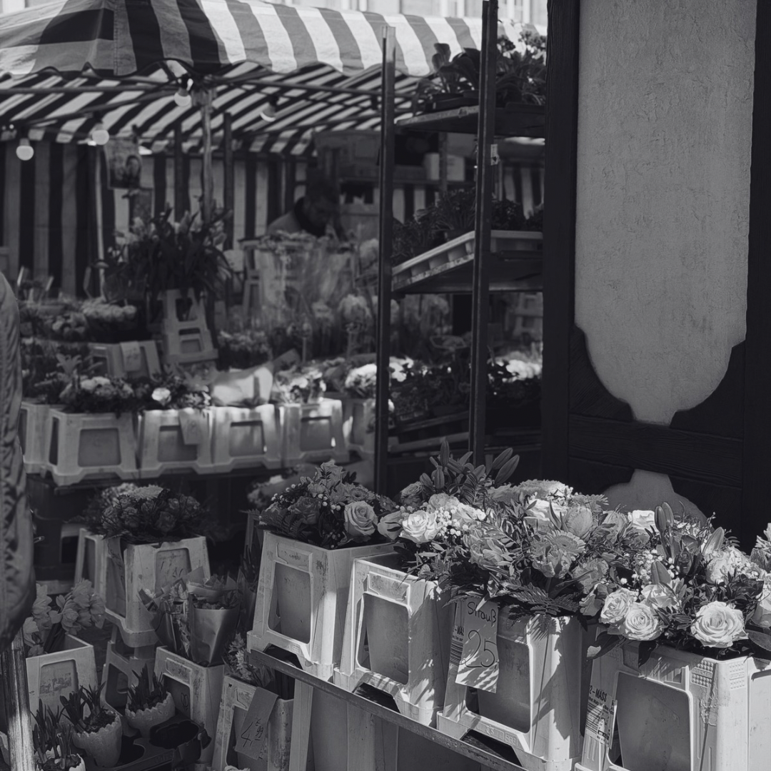 Flowers displayed in containers at an outdoor market stall with striped umbrellas overhead.
