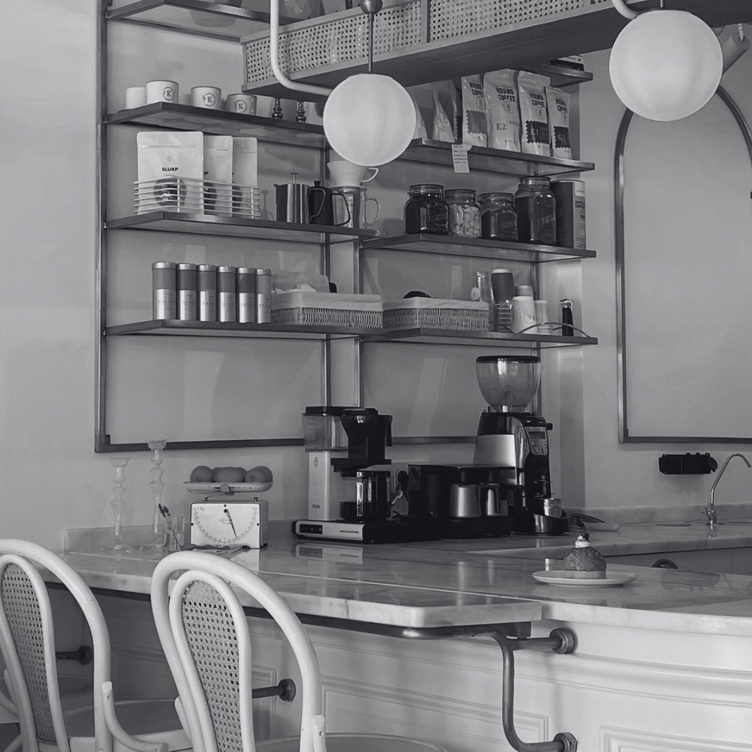 A coffee shop interior with shelves holding coffee mugs, jars, and coffee supplies, a coffee machine, blender, and various utensils on a marble counter, modern decor with hanging round white lights, and chairs in the foreground.