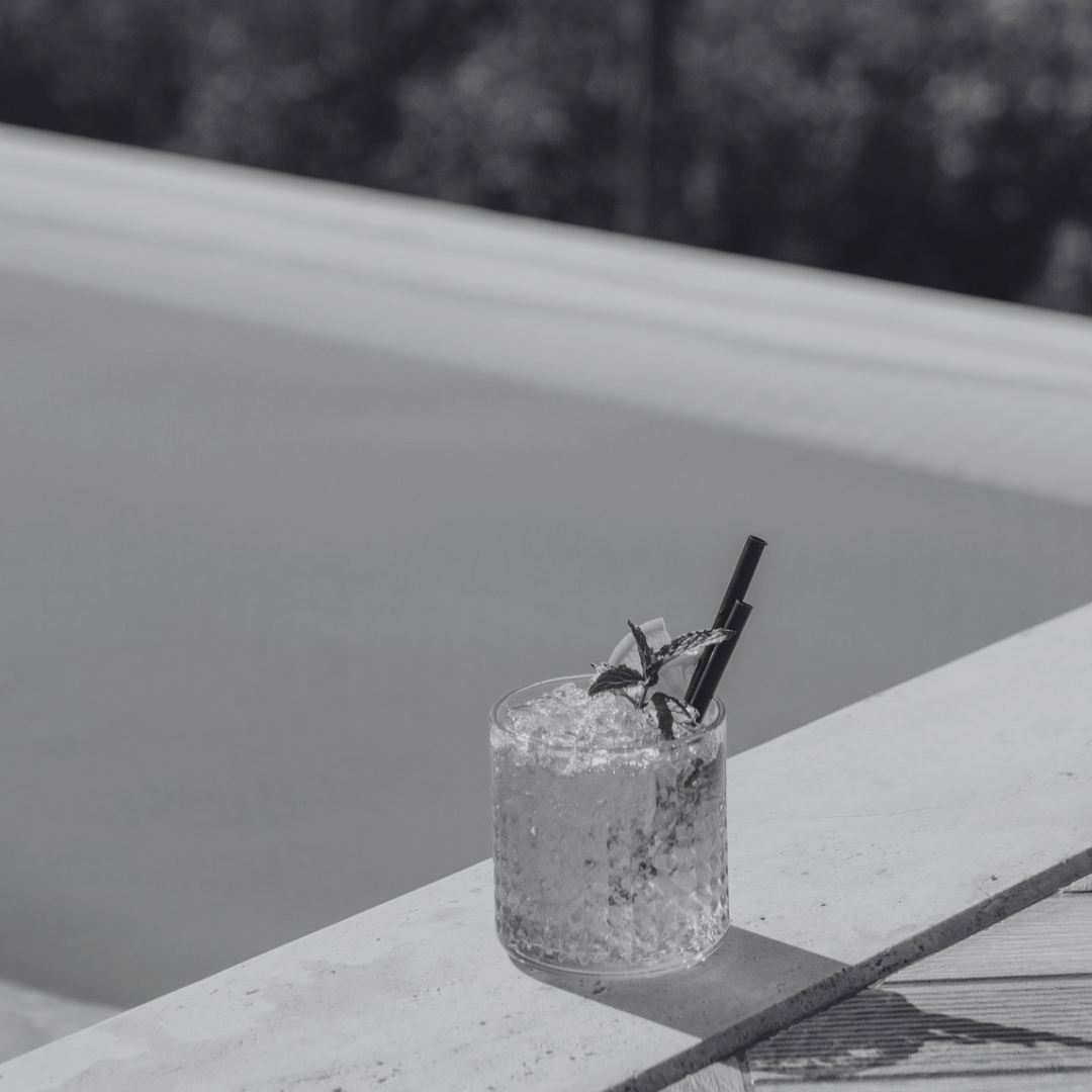 A glass of clear cocktail with ice, garnished with mint and lime, and two black straws, resting on a white ledge in black and white.