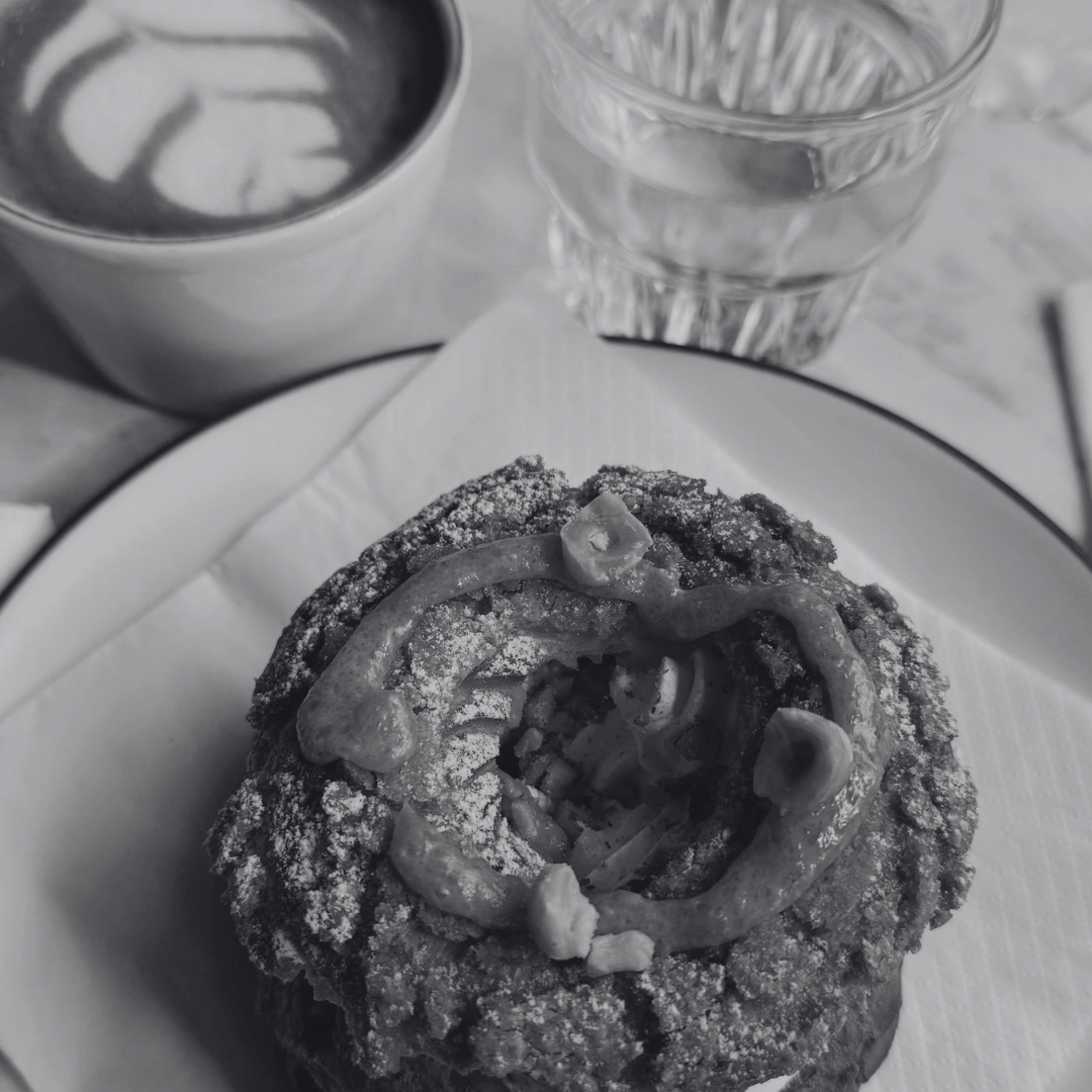 A dessert with a ring of fudge on top, dusted with powdered sugar, served on a white plate. There is a glass of water and a cup of coffee with latte art in the background.